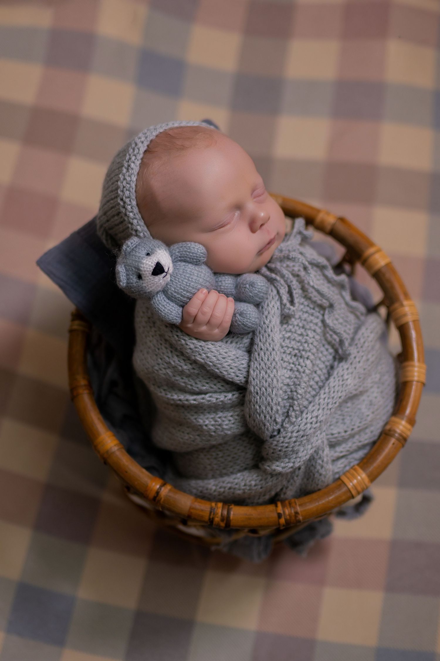 Sleeping newborn baby wrapped in gray knit blanket holding teddy bear in wicker basket on plaid background.