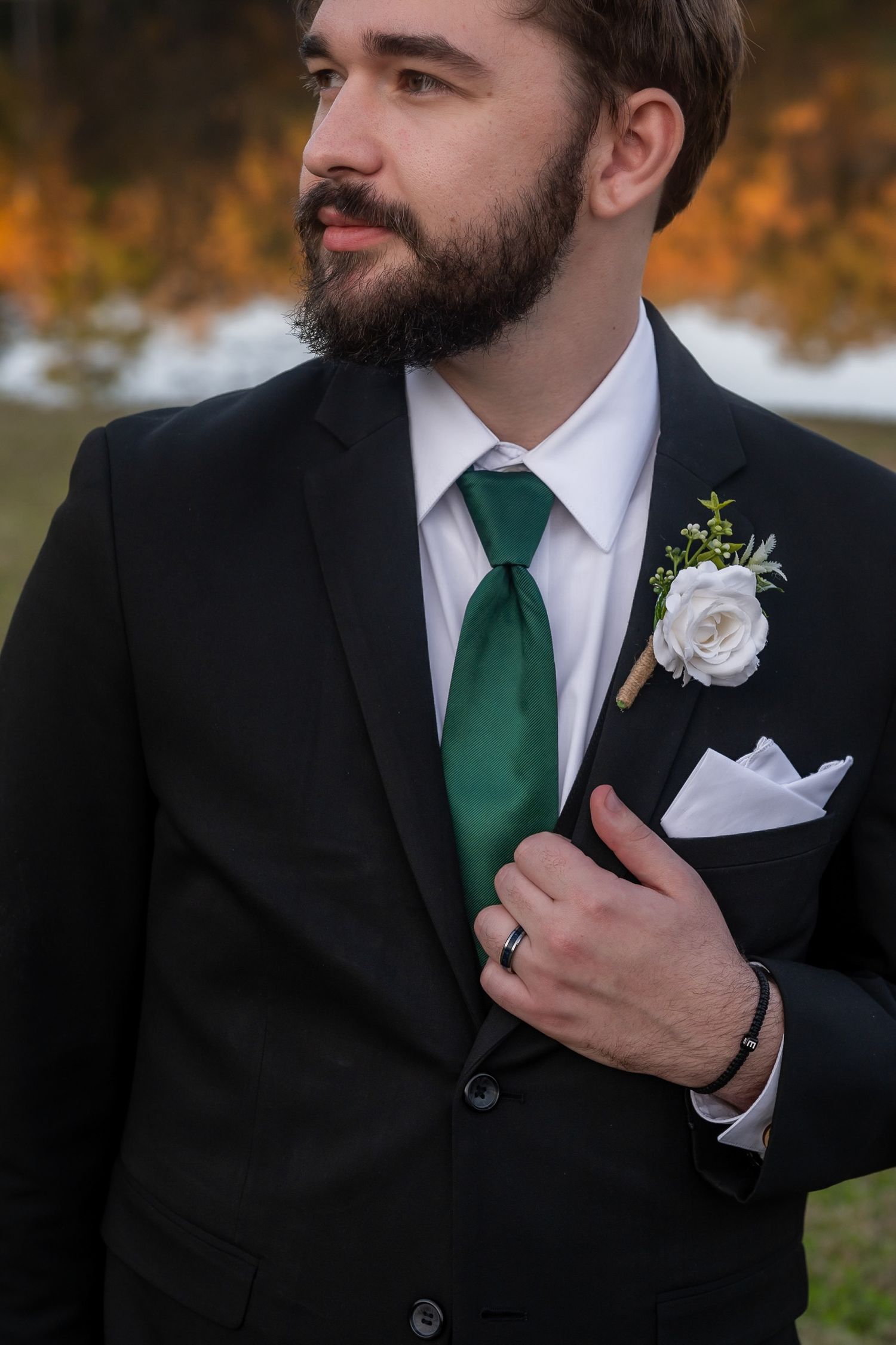 Dapper groom in black suit with green tie and white rose boutonniere posing outdoors near autumn lake.