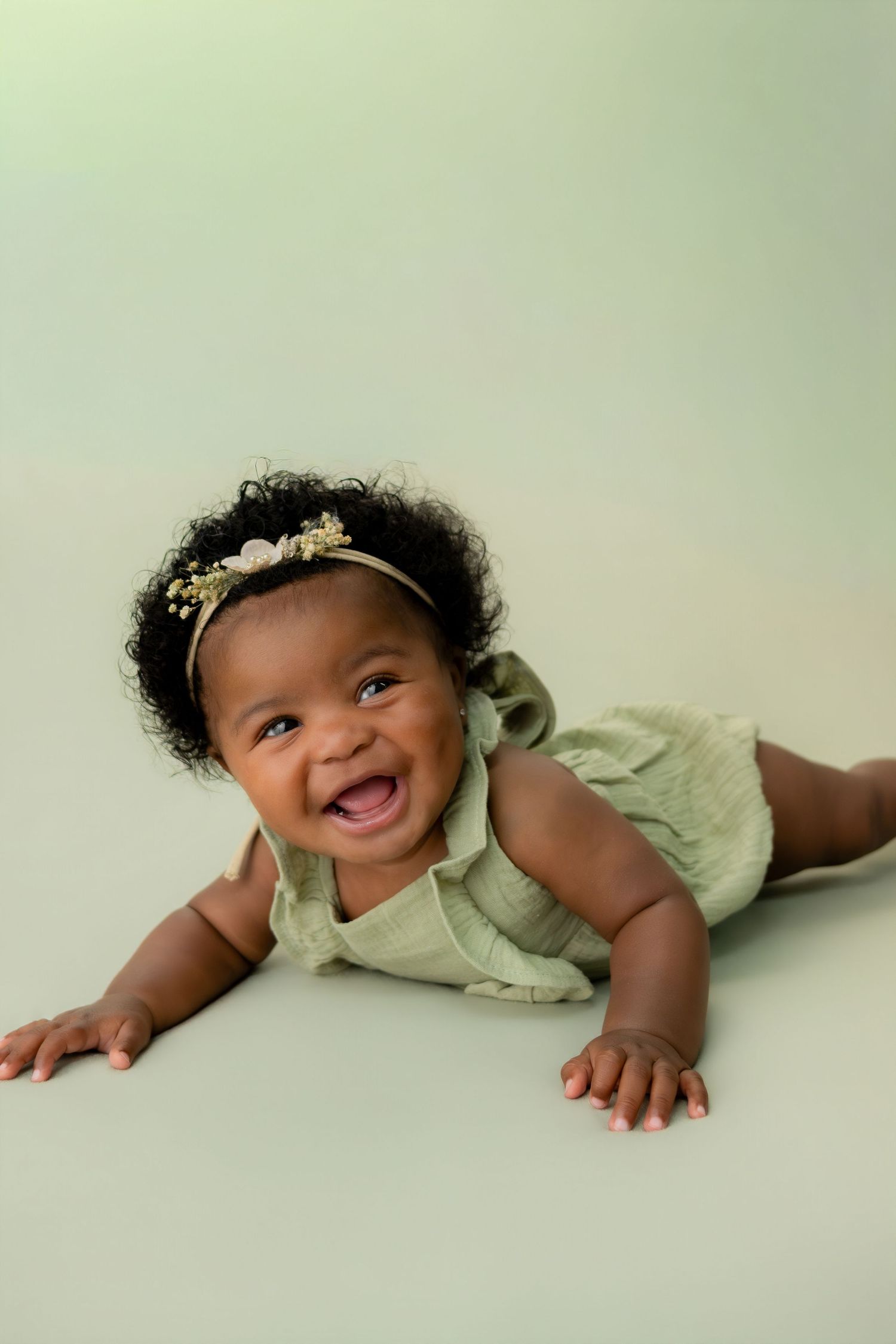 Smiling baby girl in green romper and floral headband doing tummy time on a soft green background.