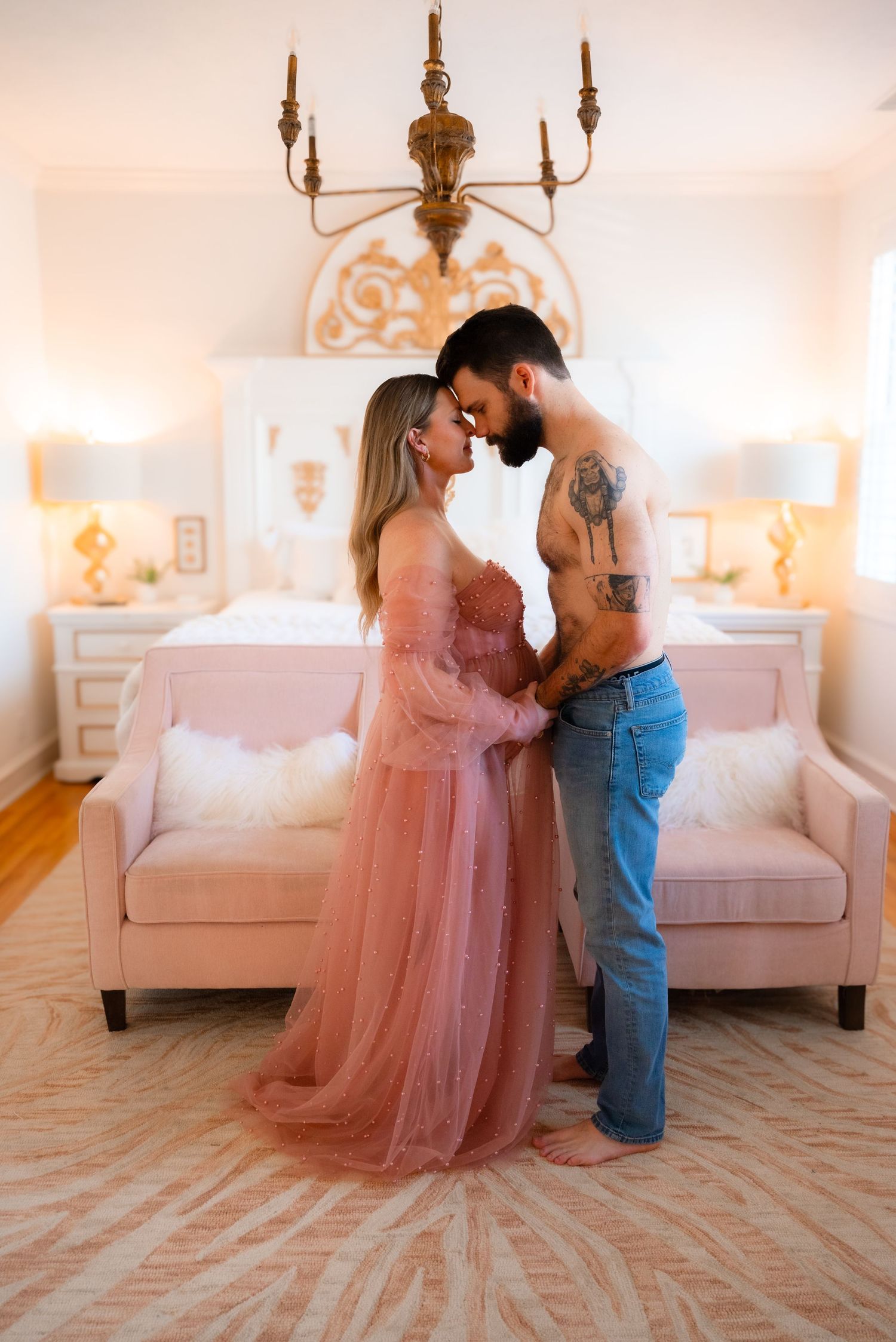 Maternity couple in pink tulle gown and jeans sharing intimate moment in elegant bedroom.