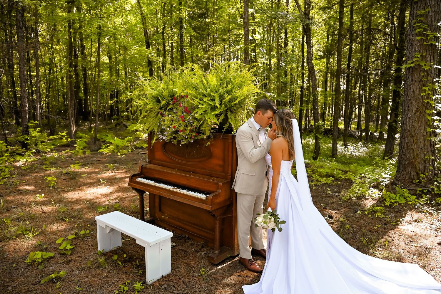 Bride and groom share a romantic kiss beside a rustic piano adorned with ferns in a lush forest setting.