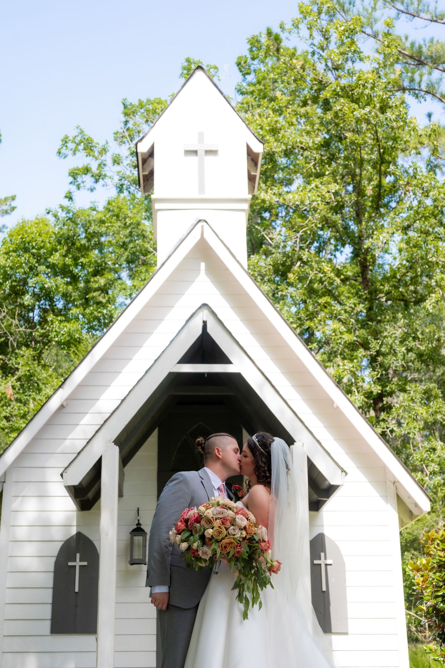 Bride and groom share a kiss in front of a charming white chapel with cross details surrounded by green trees.