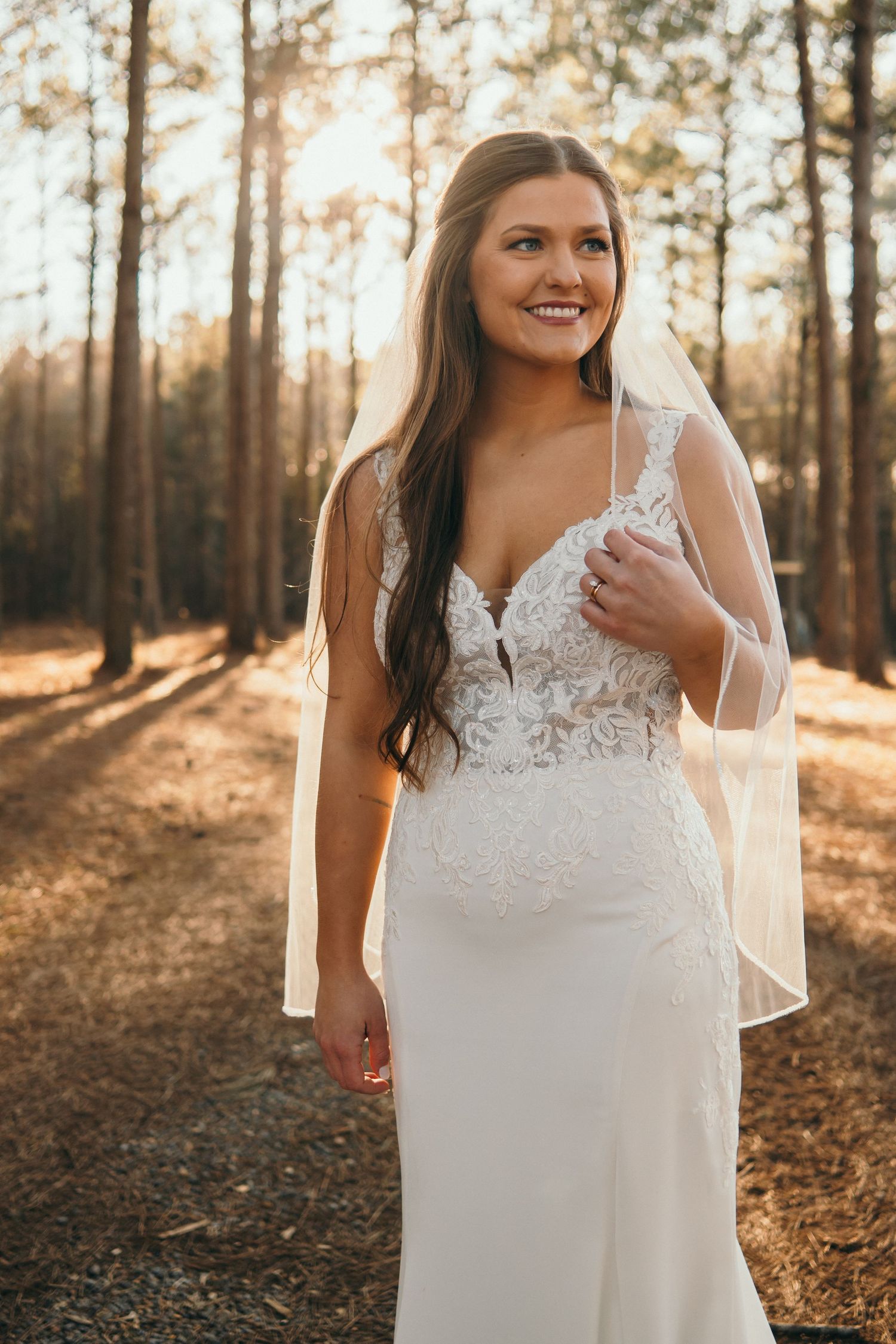 Radiant bride in lace wedding gown with veil smiling among pine trees in golden sunlight.