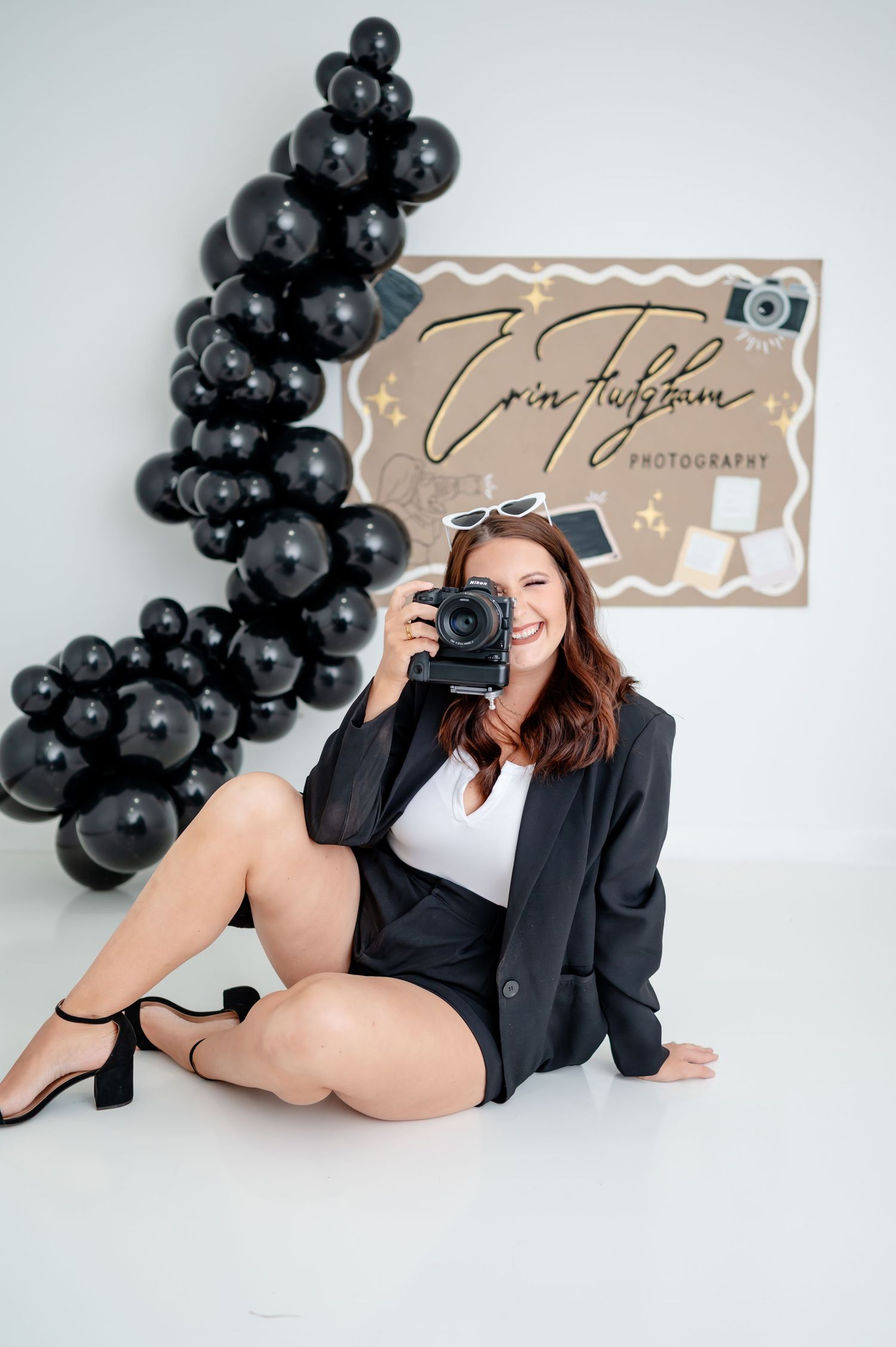 Smiling female photographer holding camera in front of black balloon arch and branded photography sign.