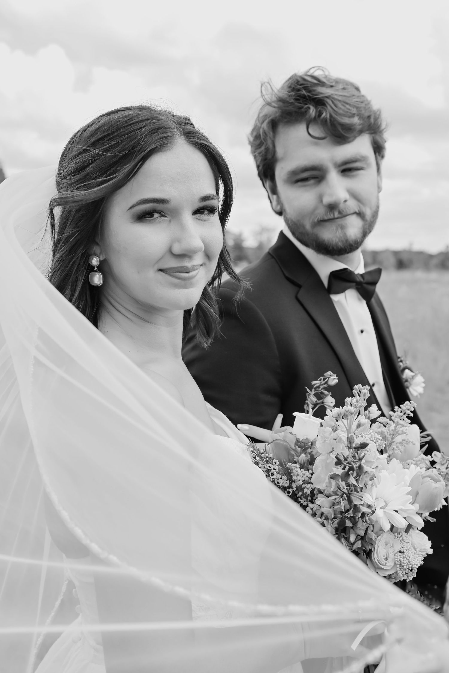 Black and white wedding portrait of smiling bride holding bouquet with groom in tuxedo behind her.