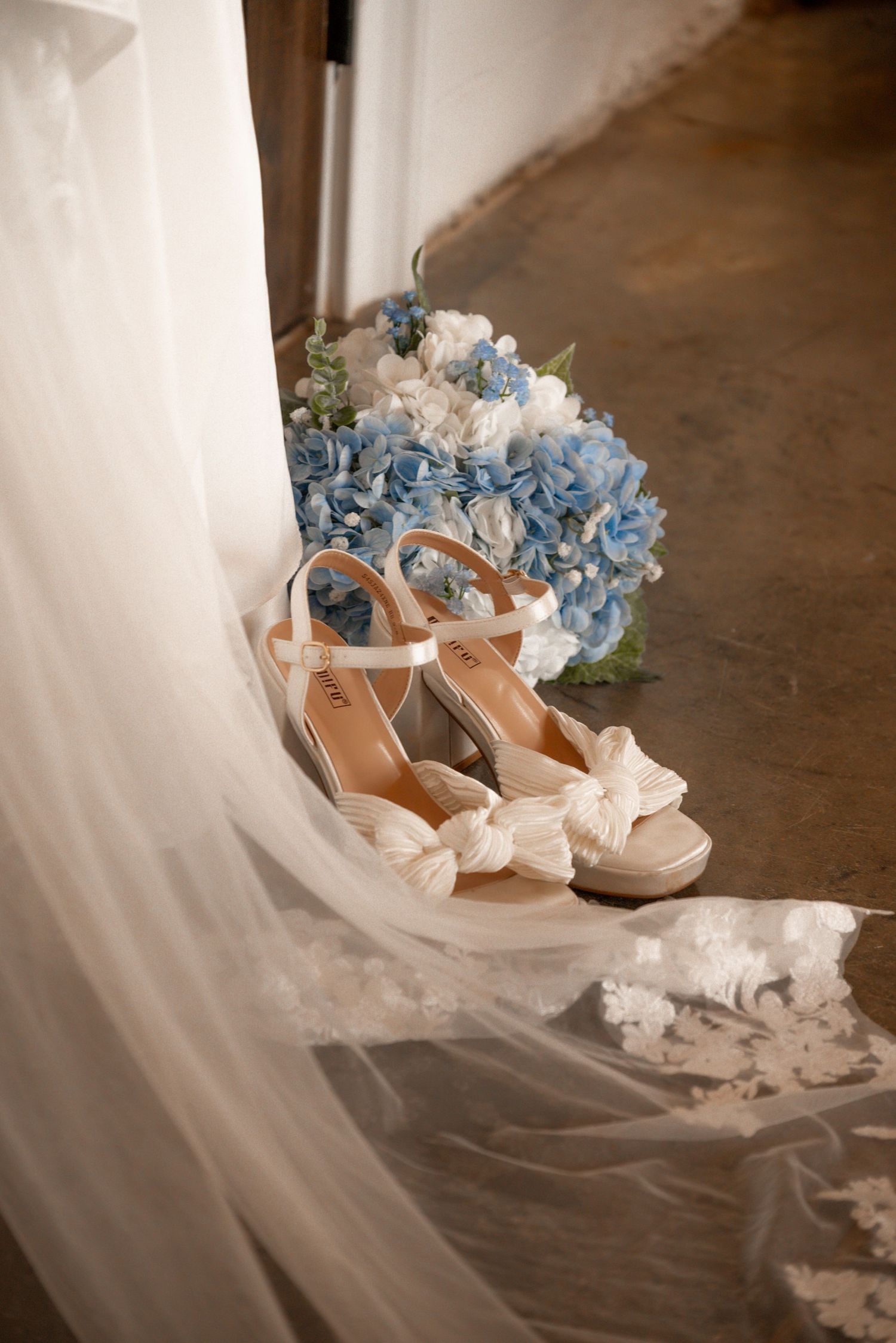 Bridal wedge heels with bow detail resting on veil beside blue and white hydrangea bouquet.