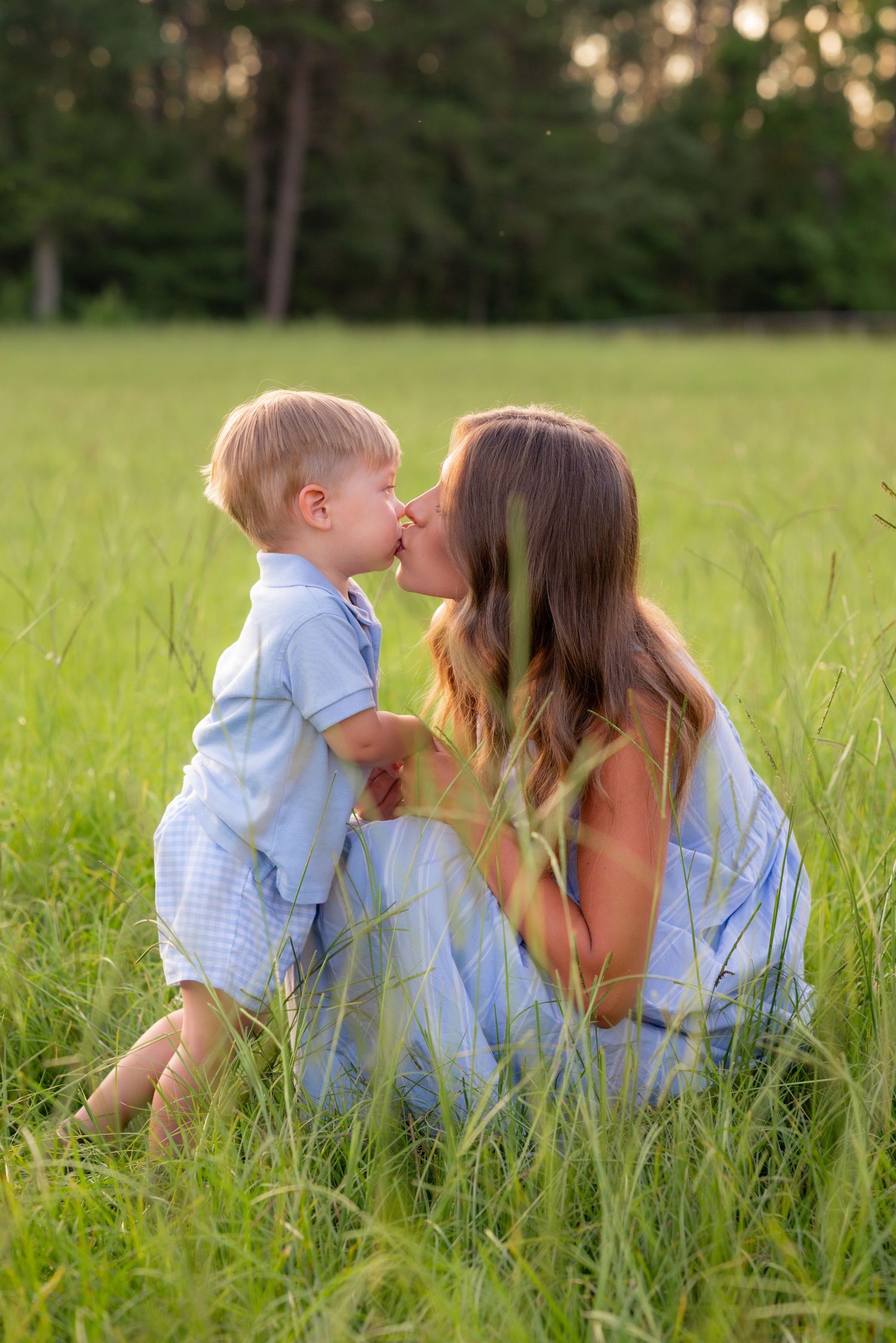 Mother and toddler son sharing a kiss while sitting in tall green grass field during golden hour sunset.