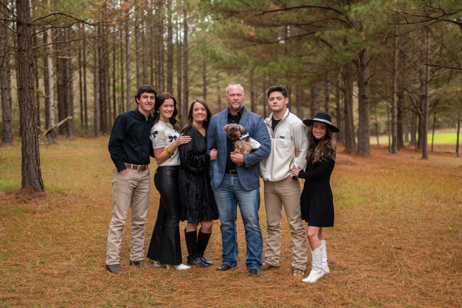 Six family members smile together holding a small dog in a pine forest during an autumn photo session.