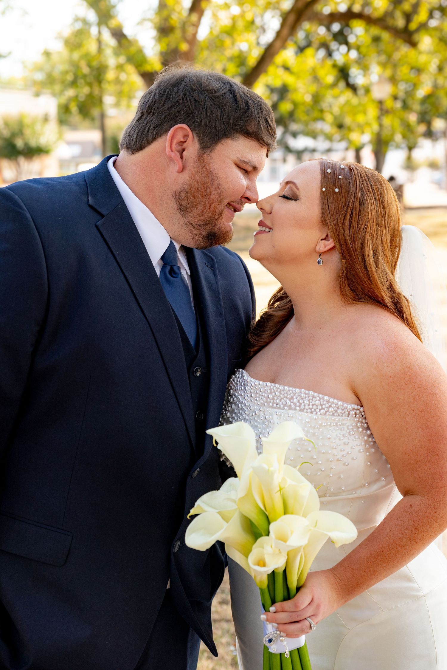 Bride and groom sharing a romantic kiss, she holds white calla lily bouquet, he wears navy suit.