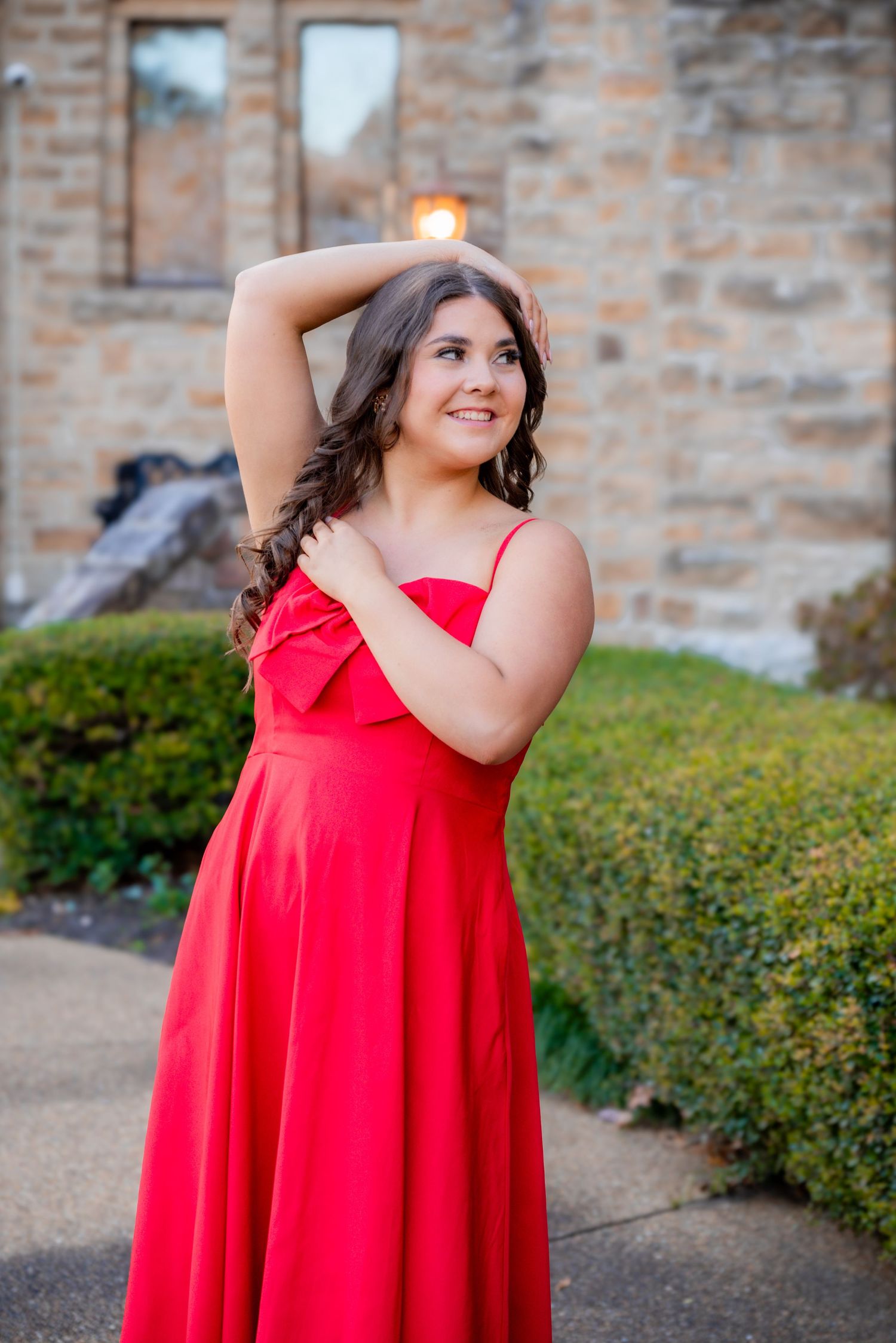 Young woman in red formal gown posing outdoors near stone building at golden hour.