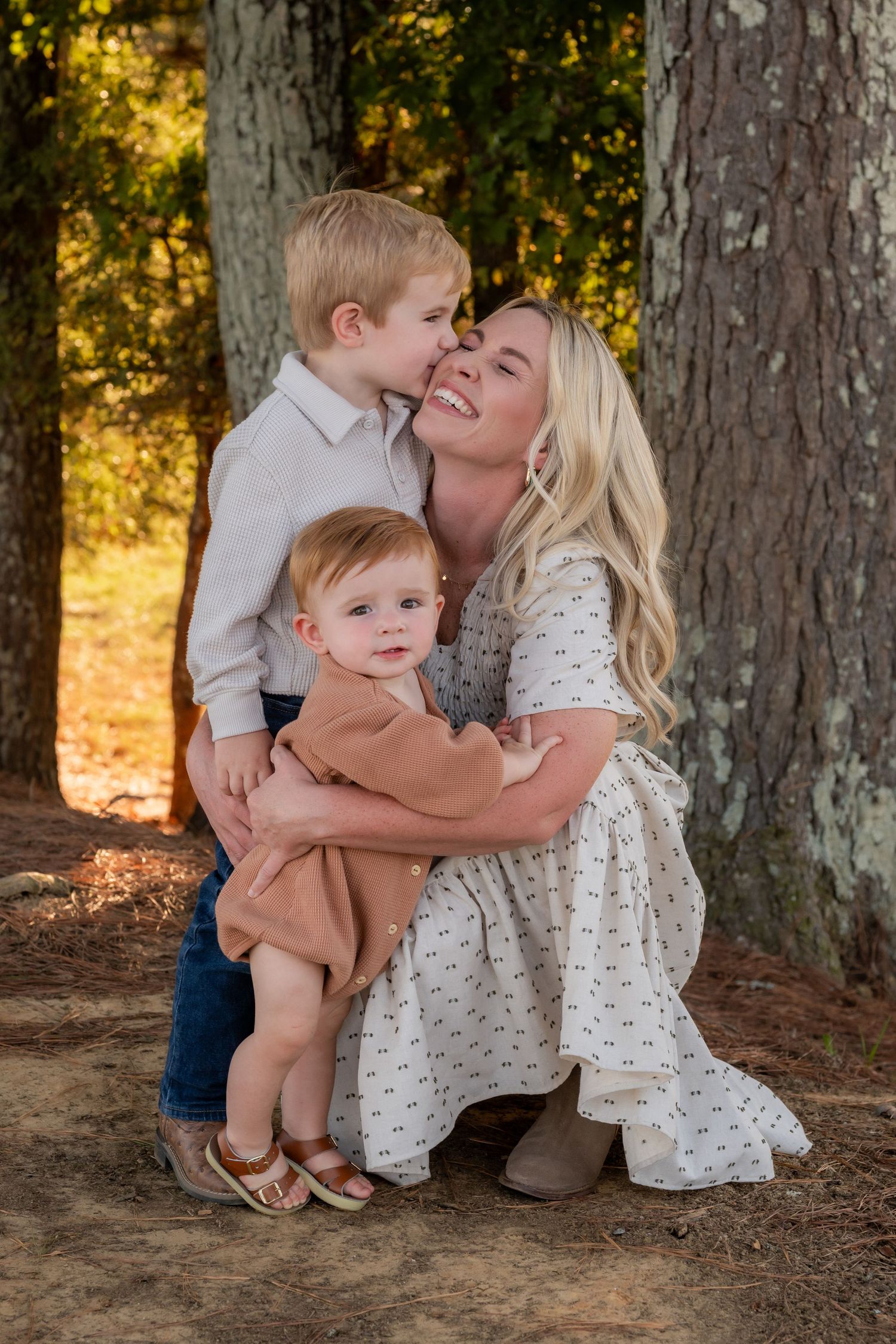 Blonde mother crouching to embrace two young children near tall pine trees in warm autumn light.