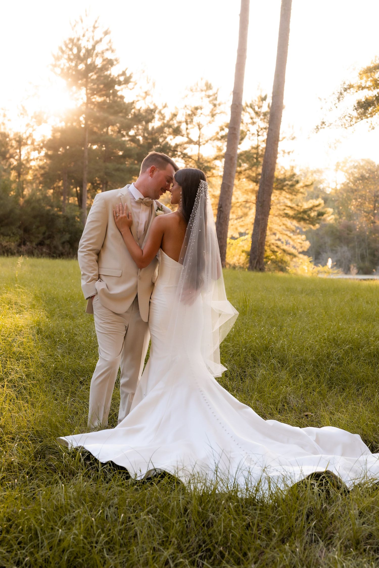 Bride and groom share a romantic kiss at golden hour in a wooded outdoor setting, bride wearing a long white gown with veil.
