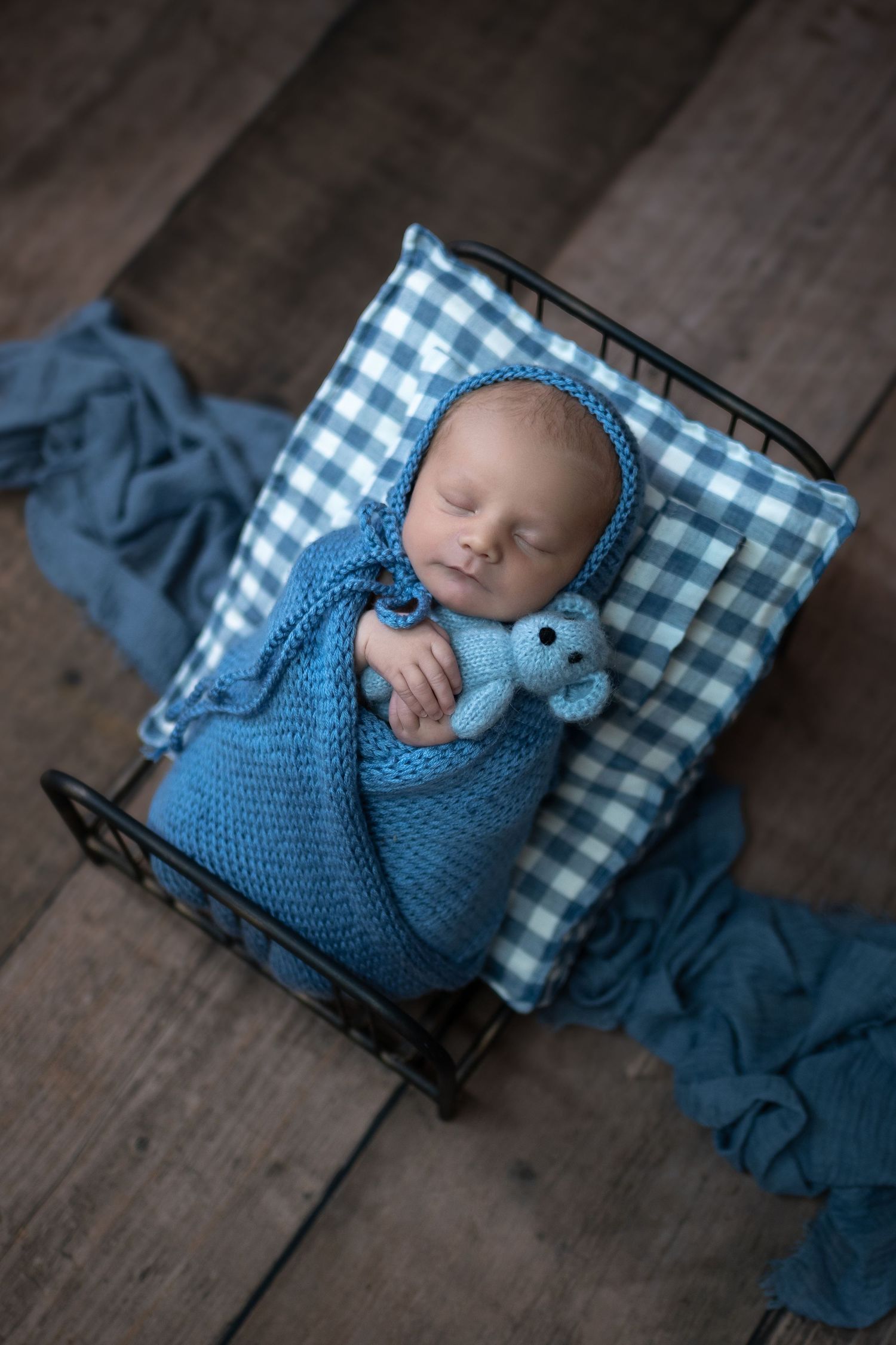 Sleeping newborn baby wrapped in blue knit swaddle with matching bonnet and teddy bear in tiny metal bed.