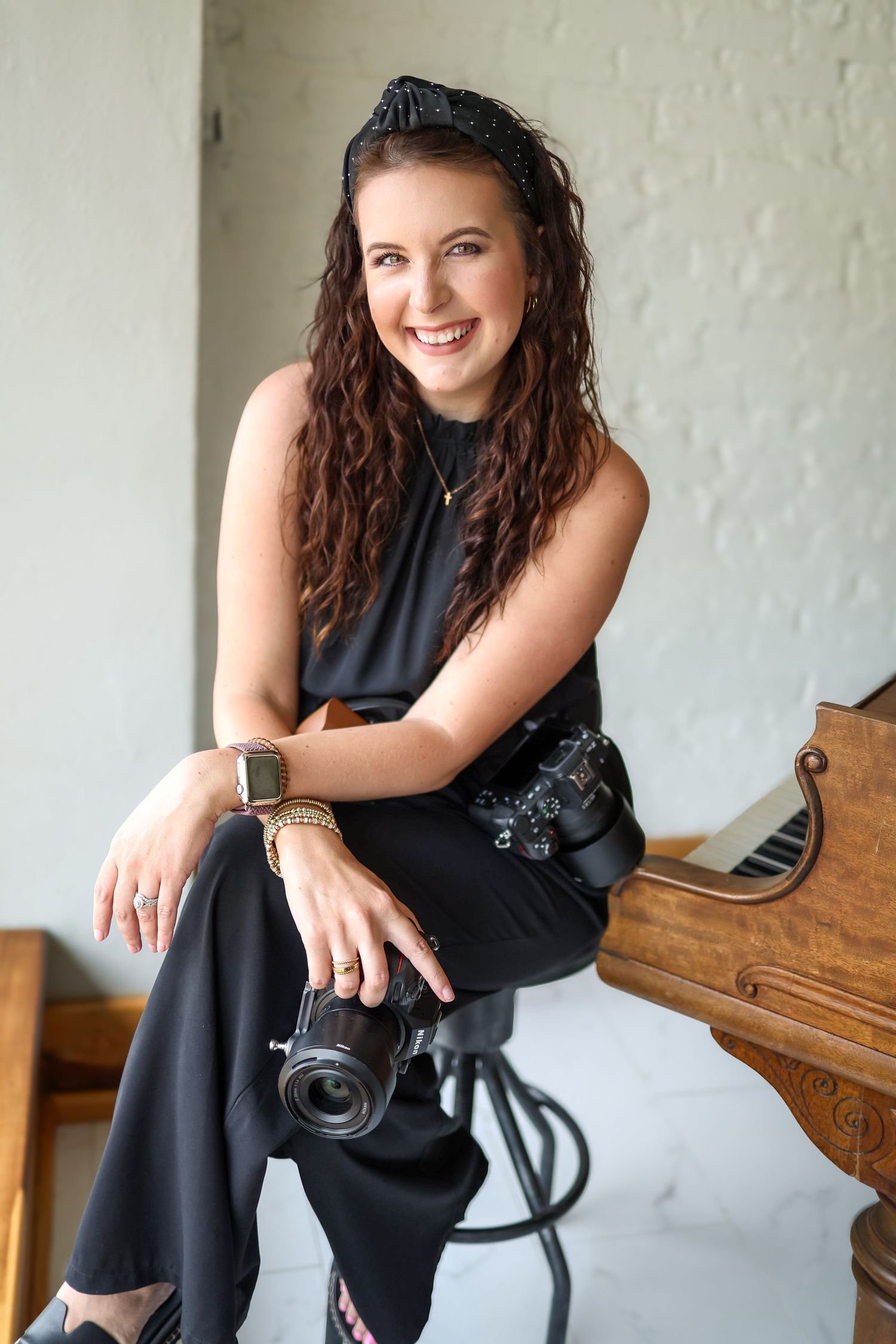 Smiling woman with curly hair sits on stool holding camera, wearing black outfit, with piano in background.