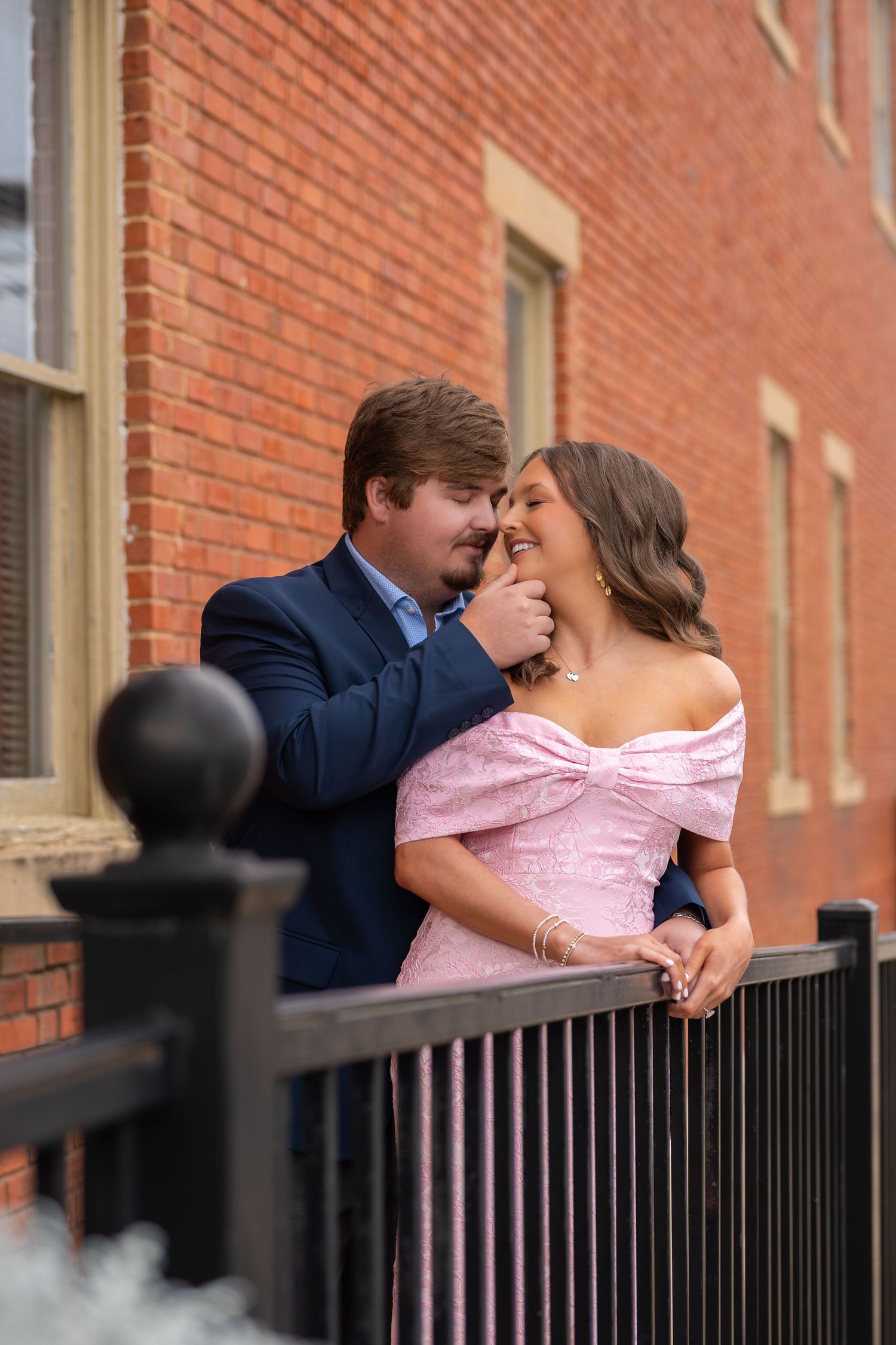 Smiling couple poses playfully against a brick building railing, woman wearing an off-shoulder pink gown.