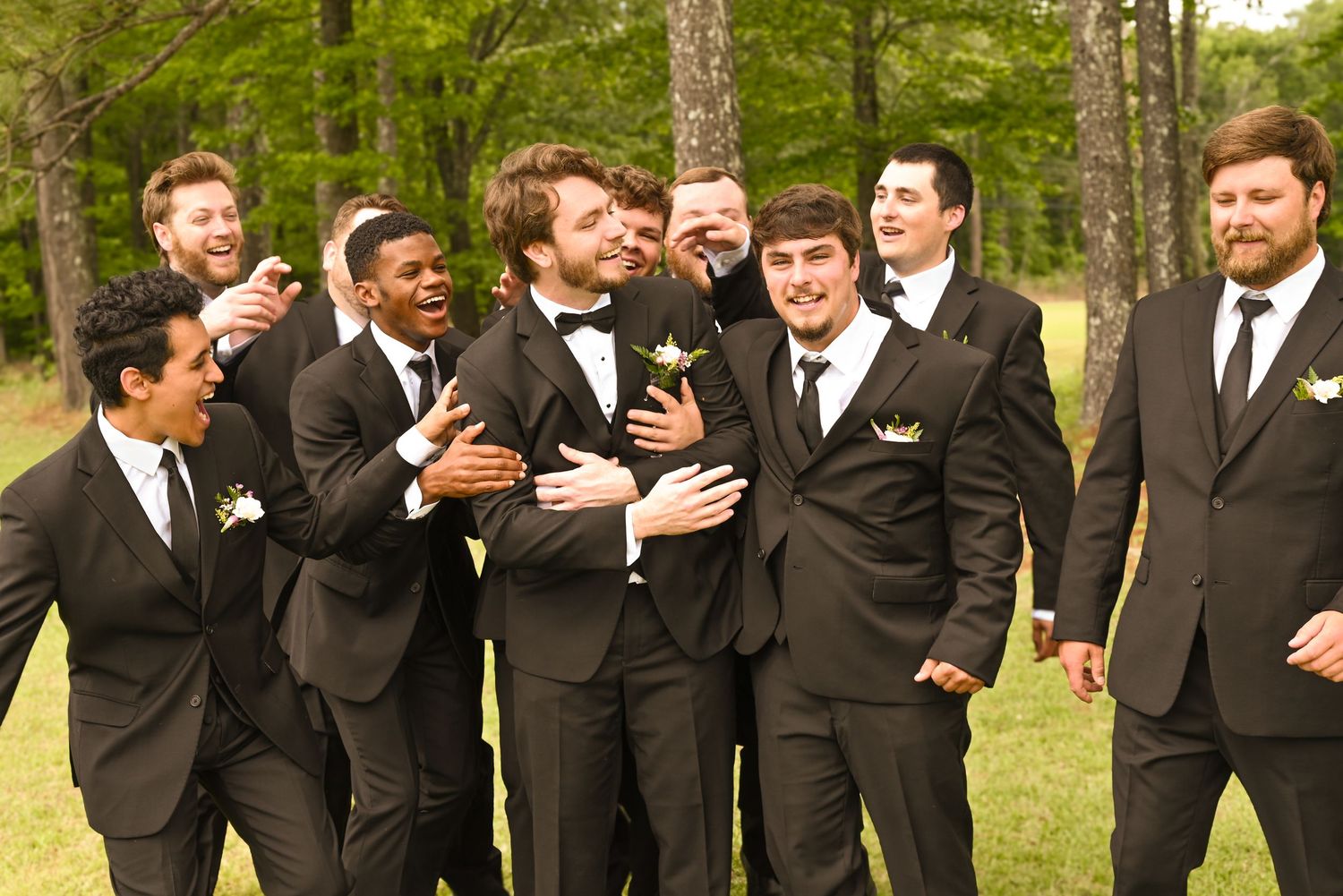 Groomsmen in black tuxedos laugh and celebrate together outdoors on a sunny wedding day.