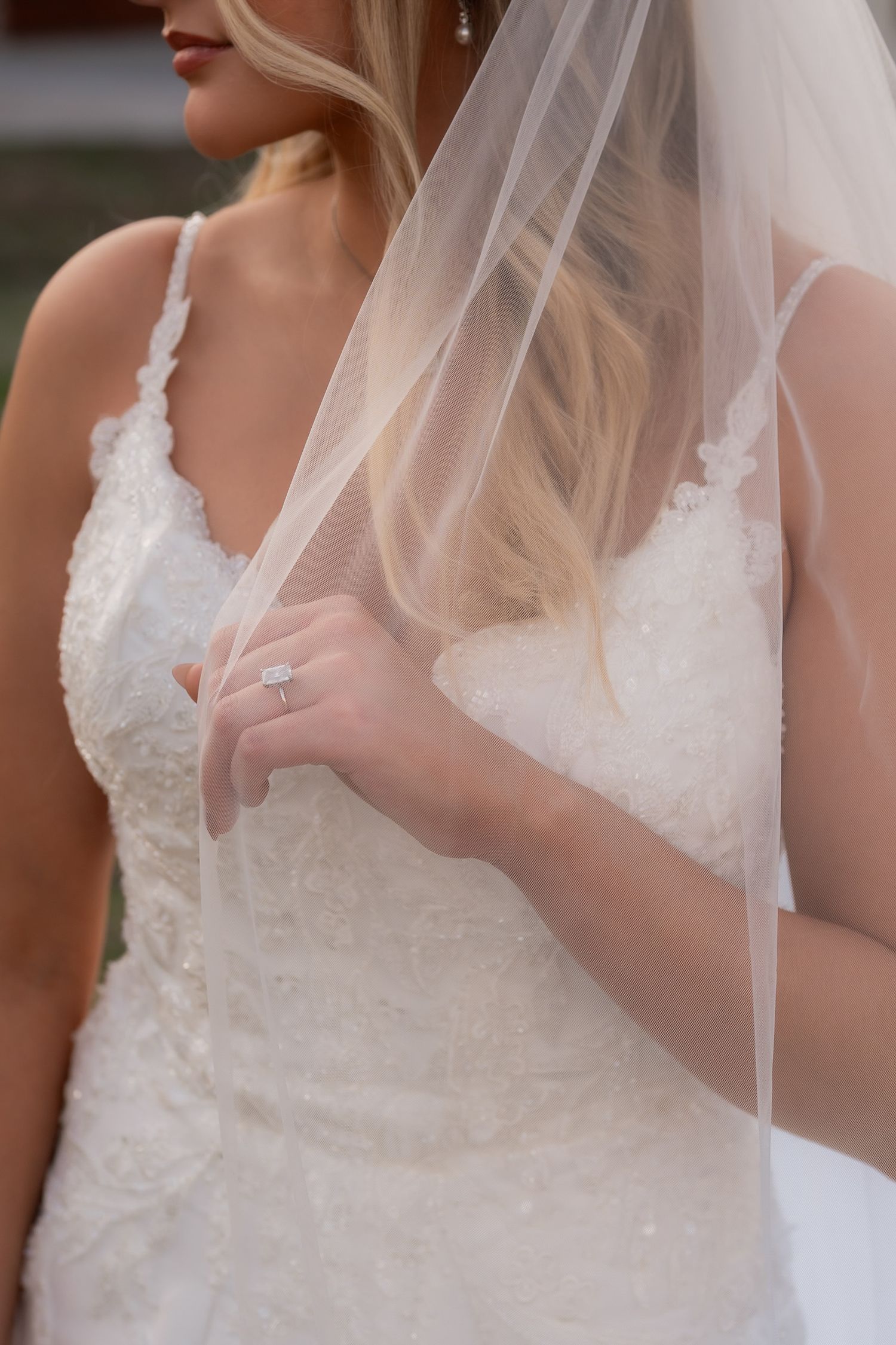 Bride in lace wedding gown holding veil, showcasing diamond engagement ring in outdoor portrait.
