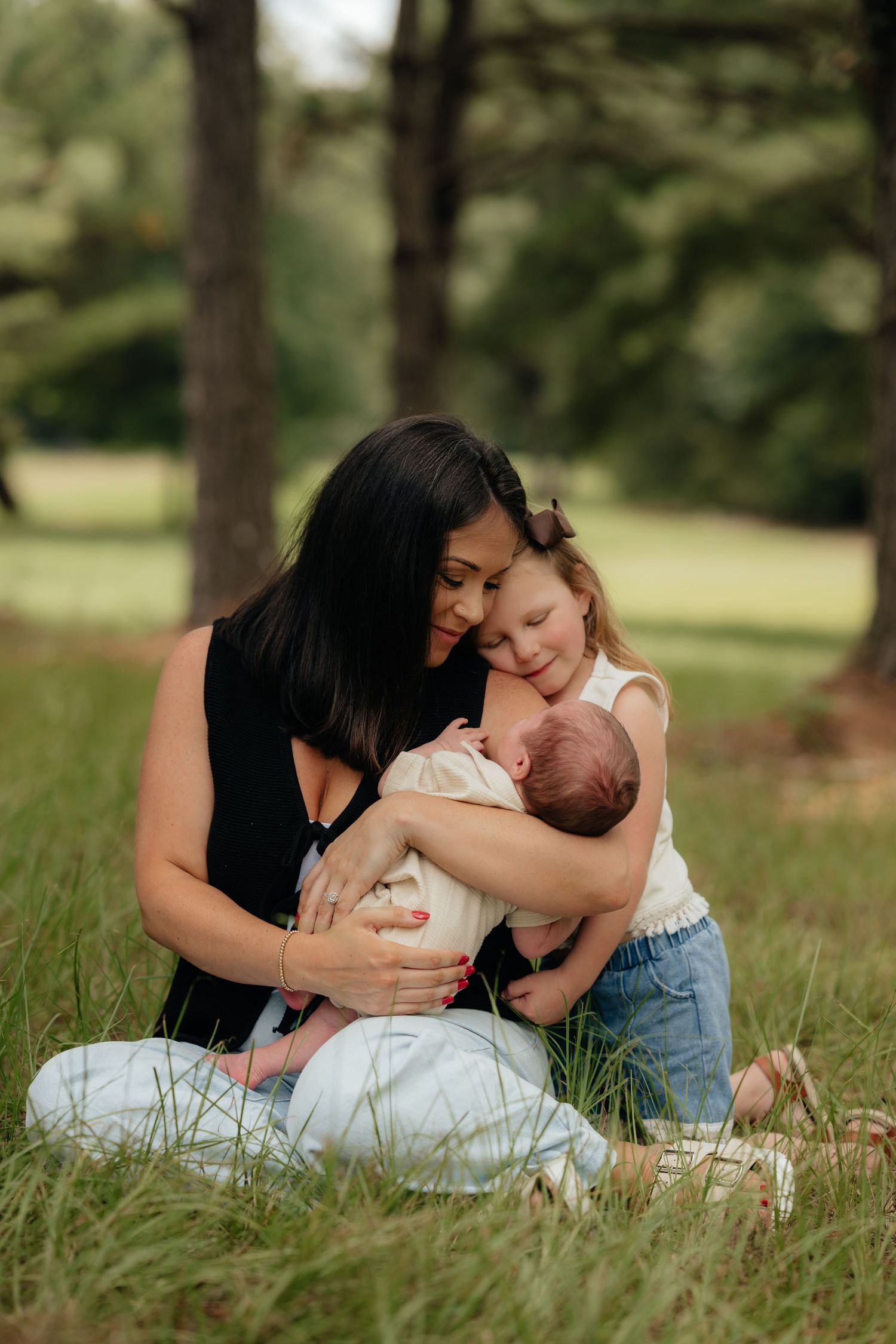 A mother sits in grass with her young daughter hugging a newborn baby in a lush green park setting.
