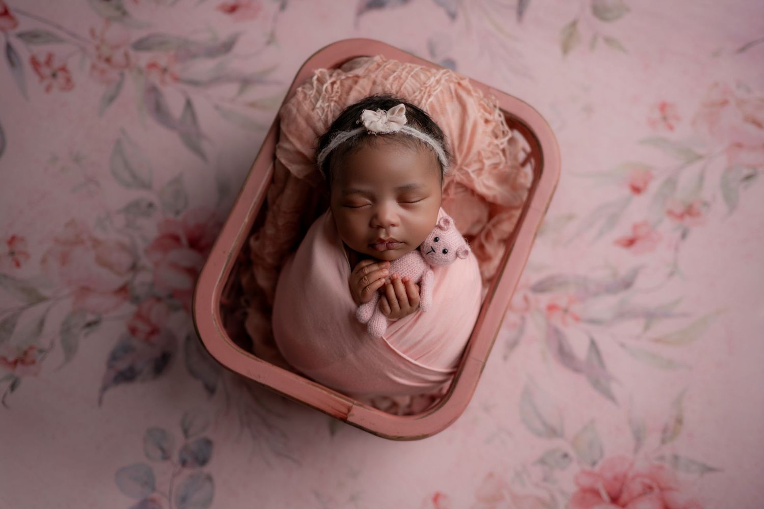 Newborn baby girl swaddled in pink wrap, sleeping in a pink basket with floral backdrop and tiny bear.
