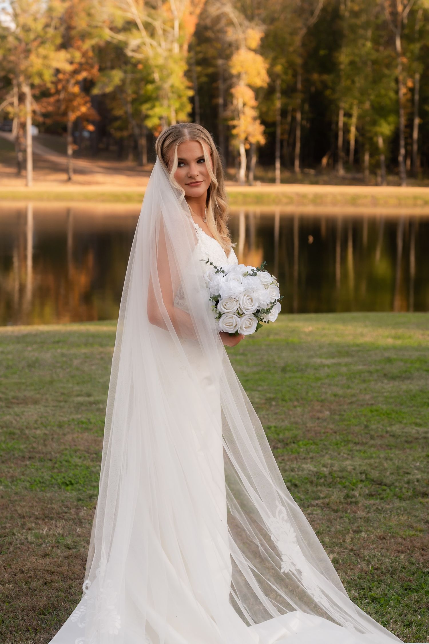 Elegant bride in flowing white gown with long veil holding white rose bouquet beside a reflective autumn lake.