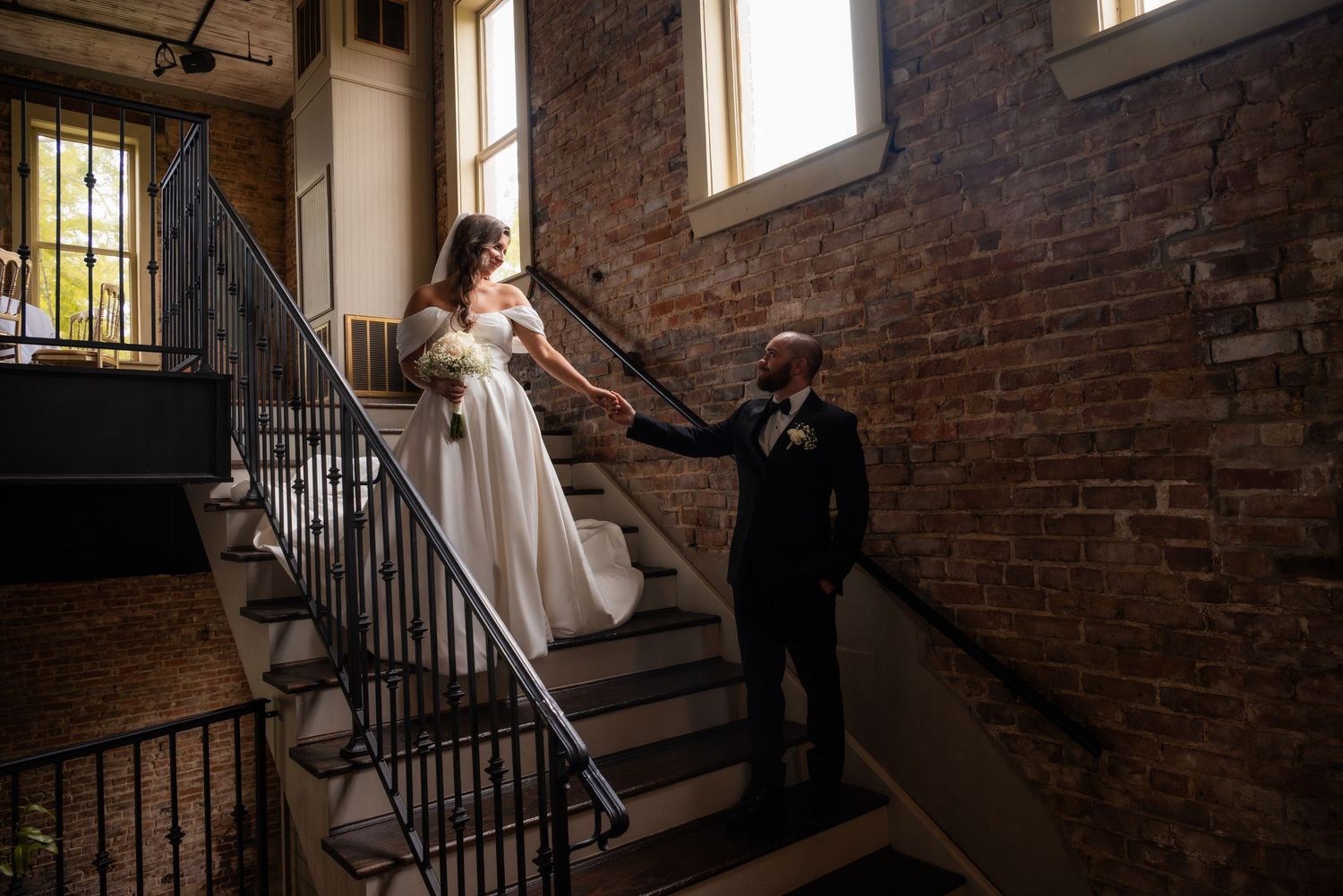 Bride and groom holding hands on staircase in rustic brick venue with natural window light.