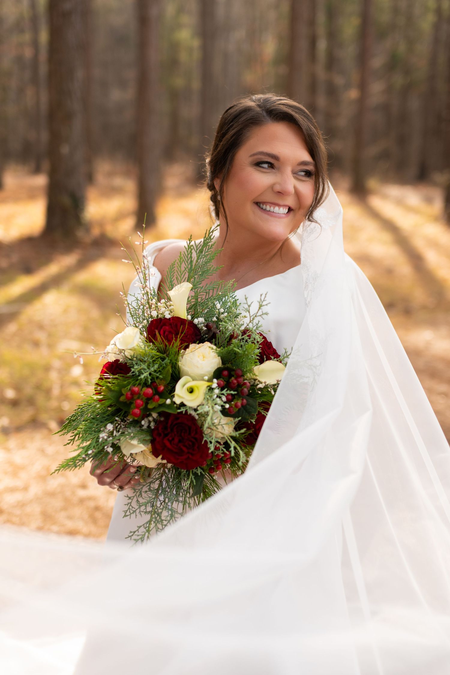 Radiant bride in white gown with flowing veil holding red and white floral bouquet in forest.
