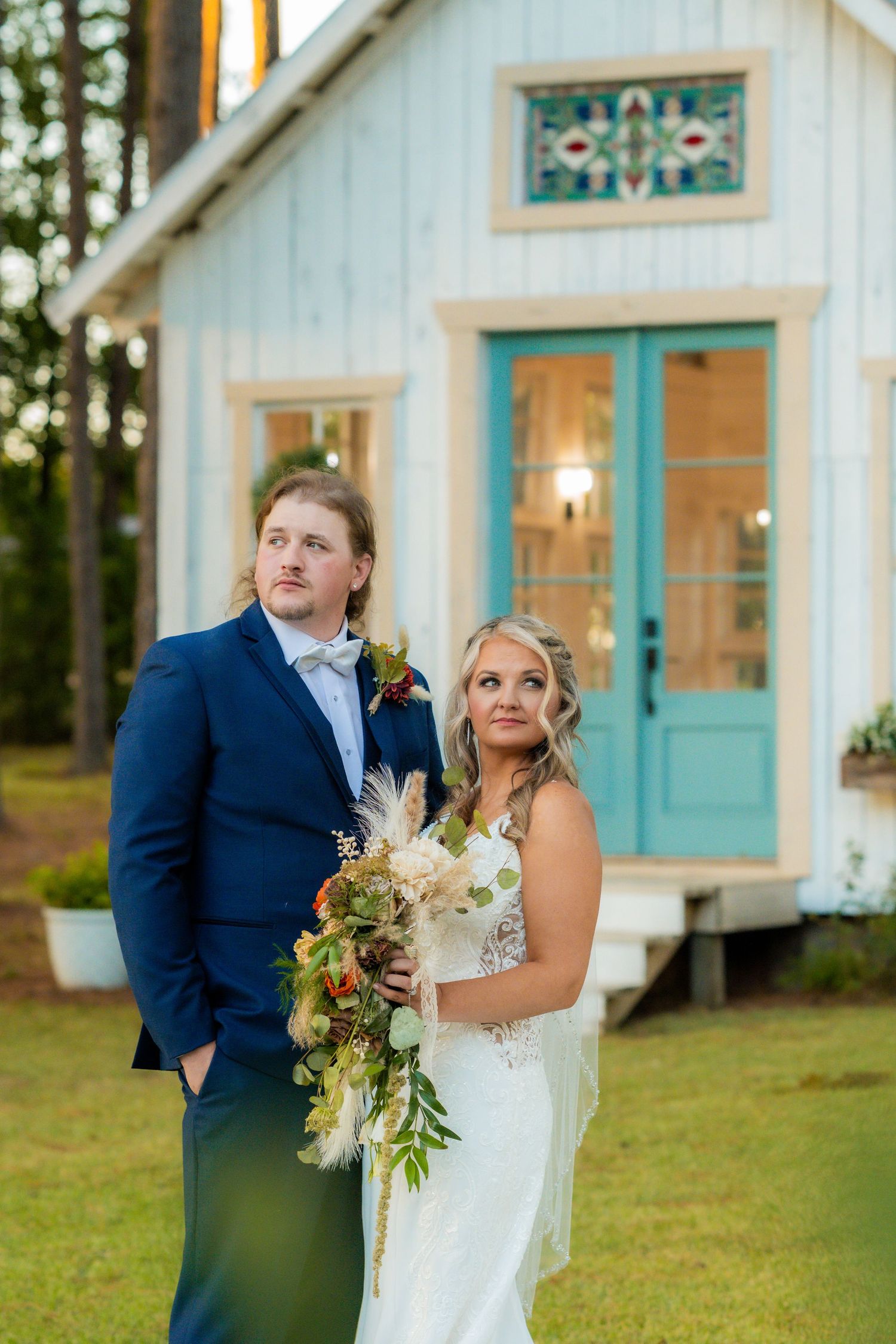 Bride and groom standing before charming white chapel with teal doors at sunset.