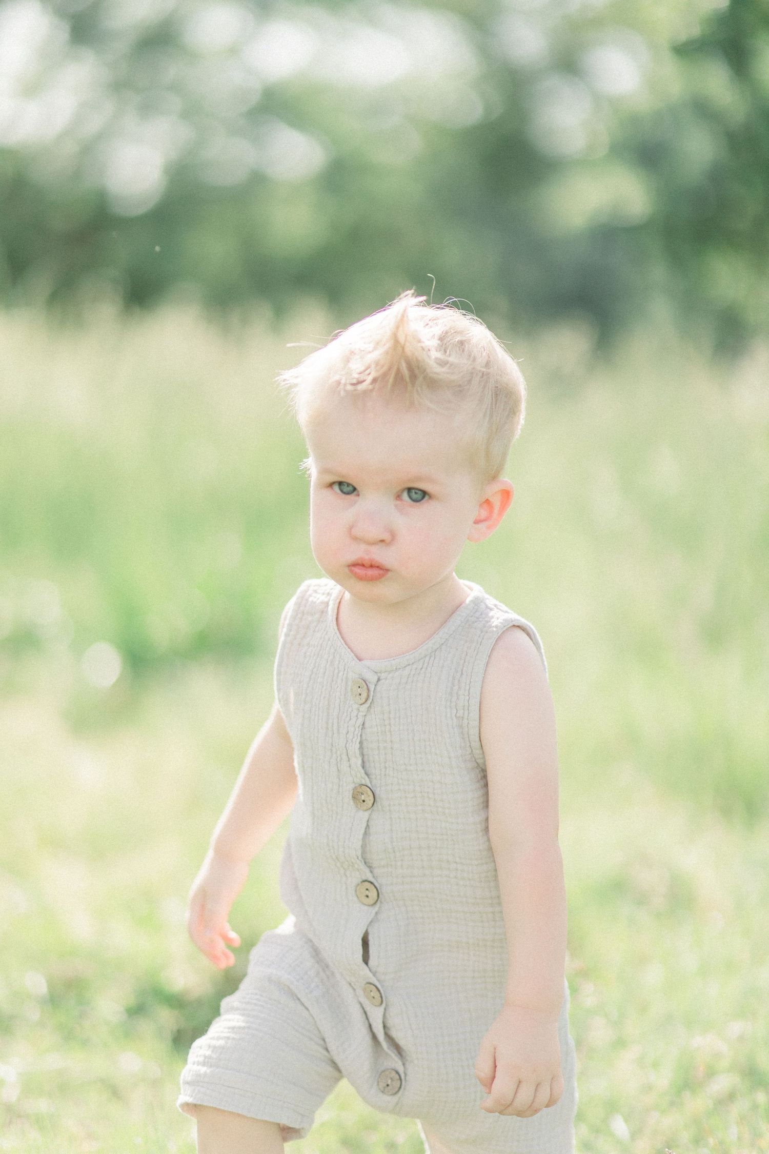 Little one wearing a beige buttoned romper standing in a sunlit summer meadow at Fellows Lake in Springfield, Missouri.