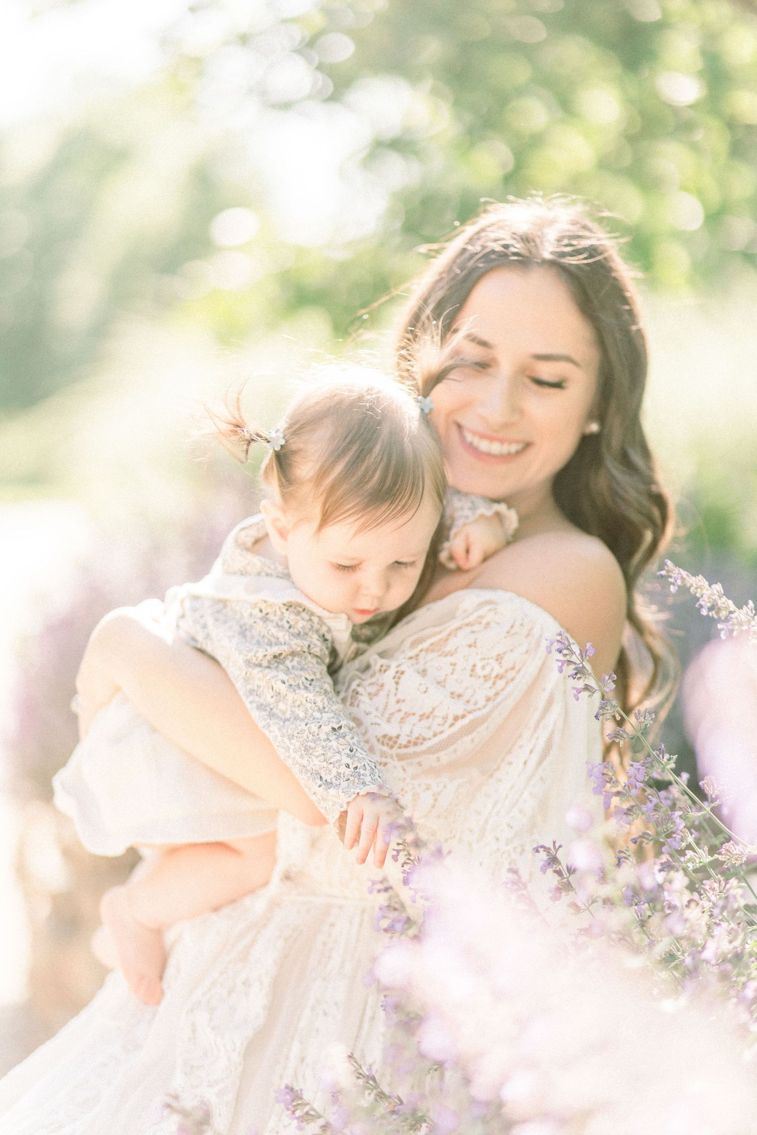 Smiling mother in lace dress holding baby girl among lavender flowers in soft sunlit garden.