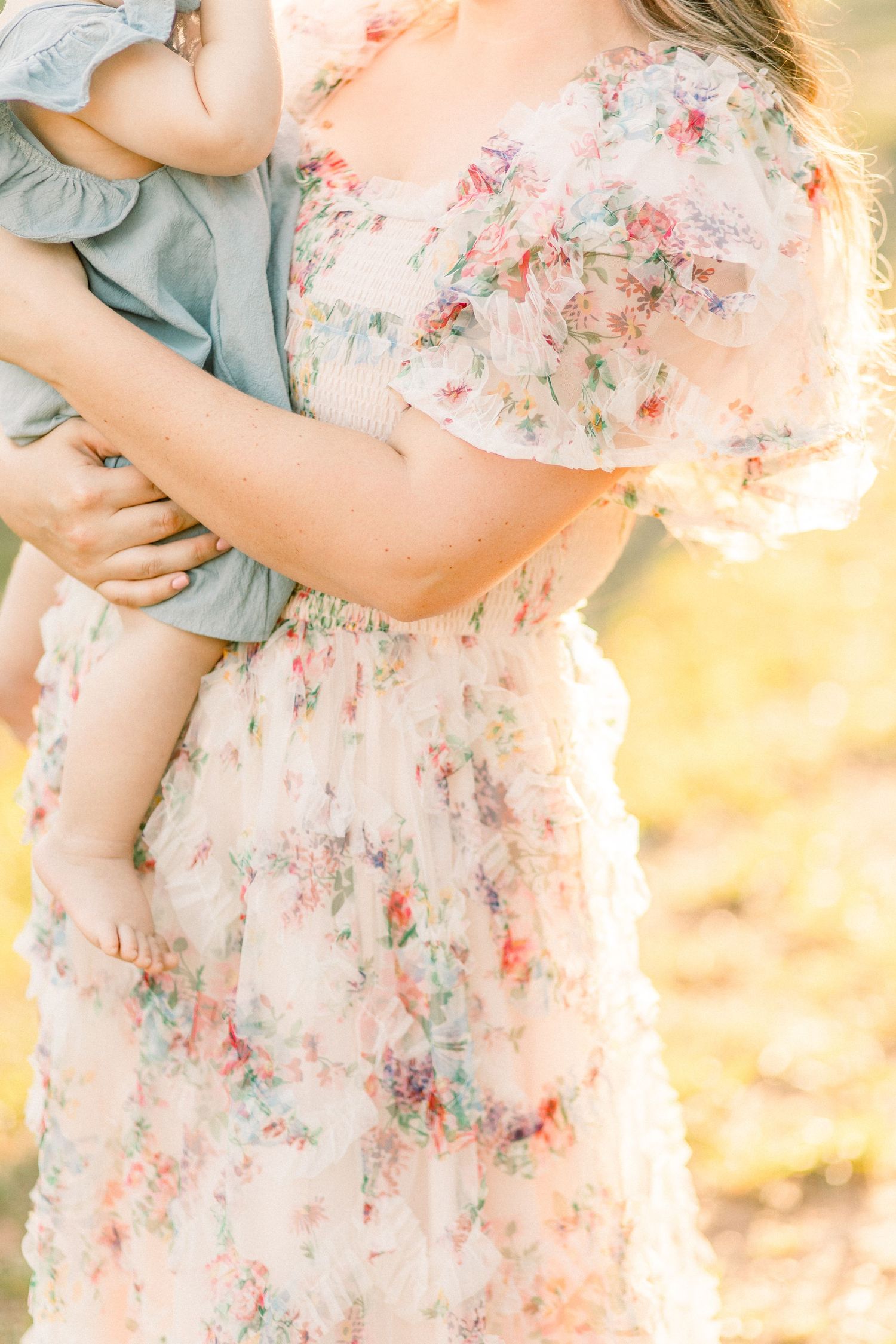 A mother in a floral dress holds her baby in a soft, sunlit outdoor setting.