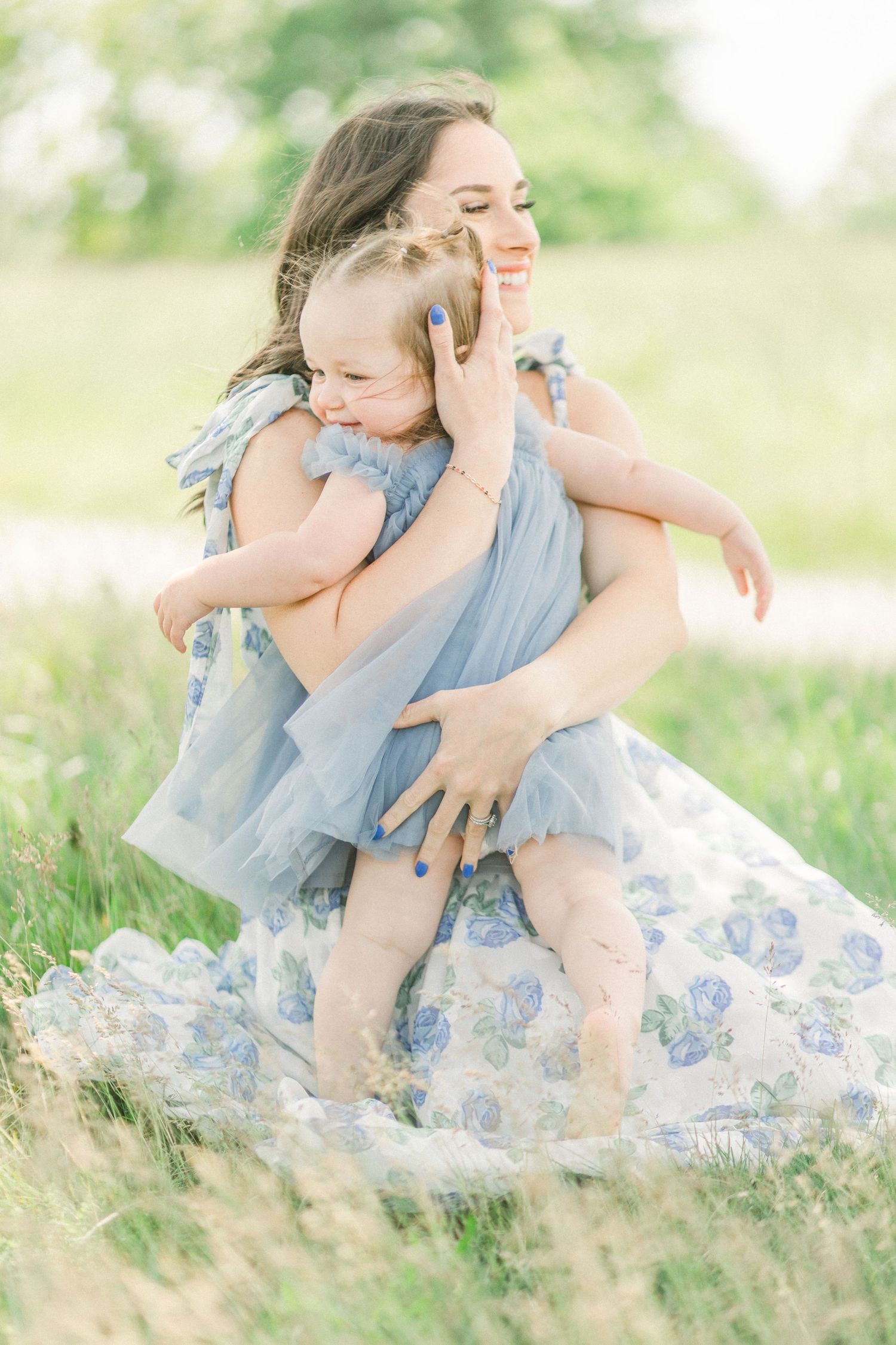 Mother in blue floral dress joyfully holds toddler daughter in matching blue tutu outdoors in green field.