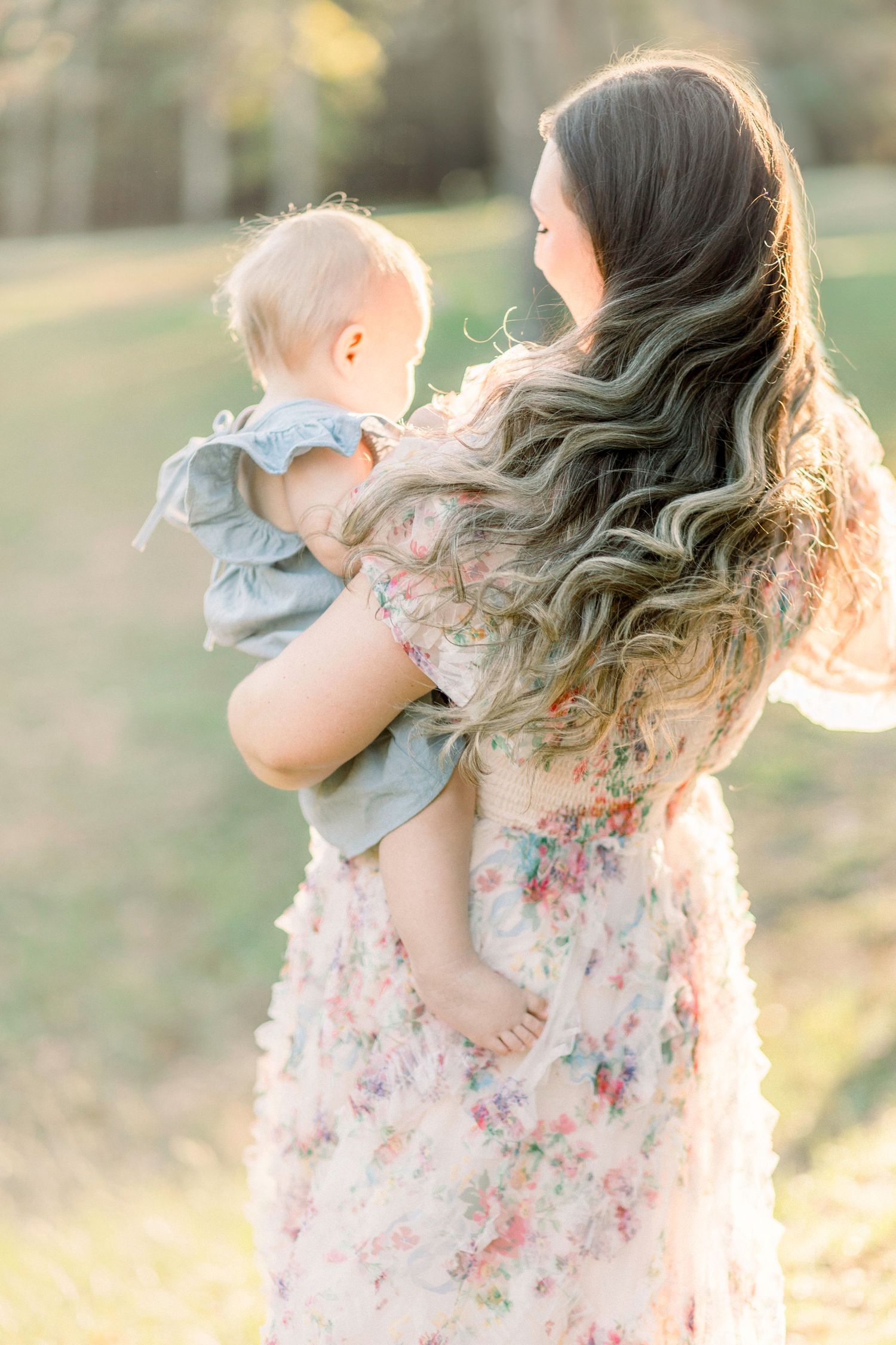 A tender moment captured in golden sunlight shows a mama in a floral dress and her baby in blue embracing in Marshfield, Missouri.