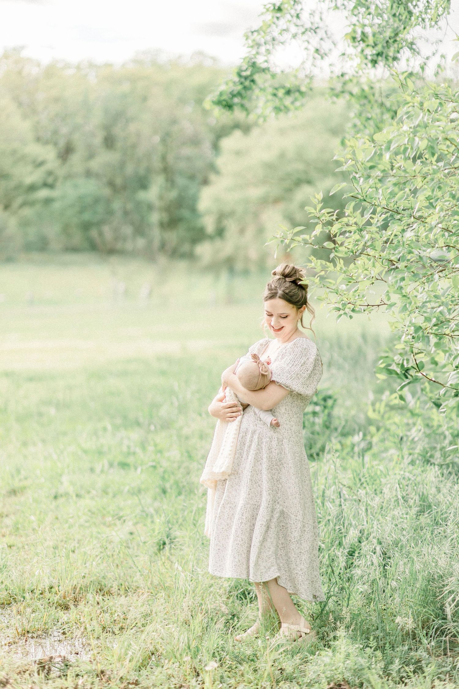 A mother in a floral dress tenderly holds her newborn baby outdoors in a lush green field.
