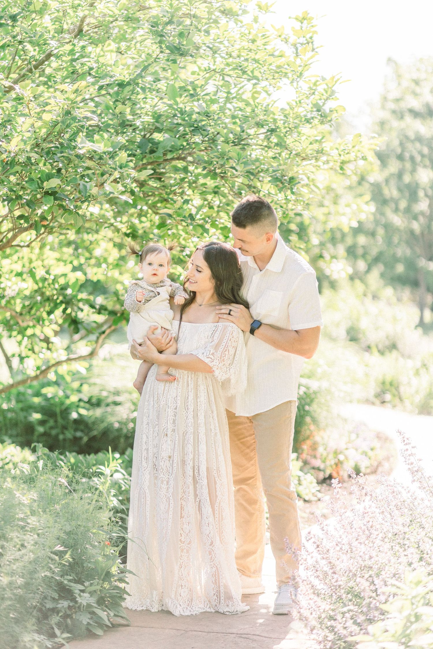 A happy family poses outdoors, mother in lace gown holding baby, father in white shirt, surrounded by lush greenery.