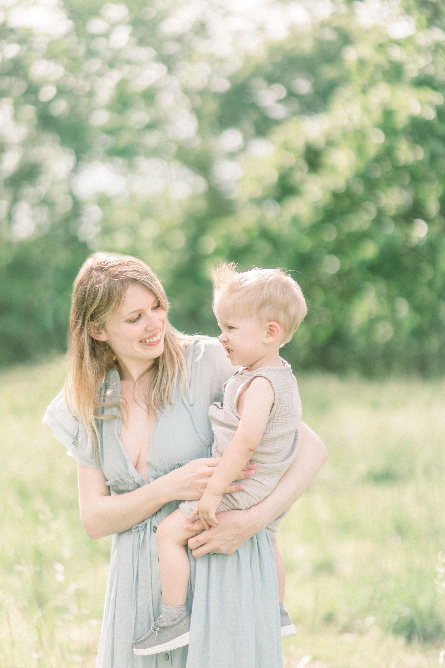 A mother in a blue dress holds her toddler while standing in a sun-dappled meadow on a bright summer day in Springfield, Missouri.