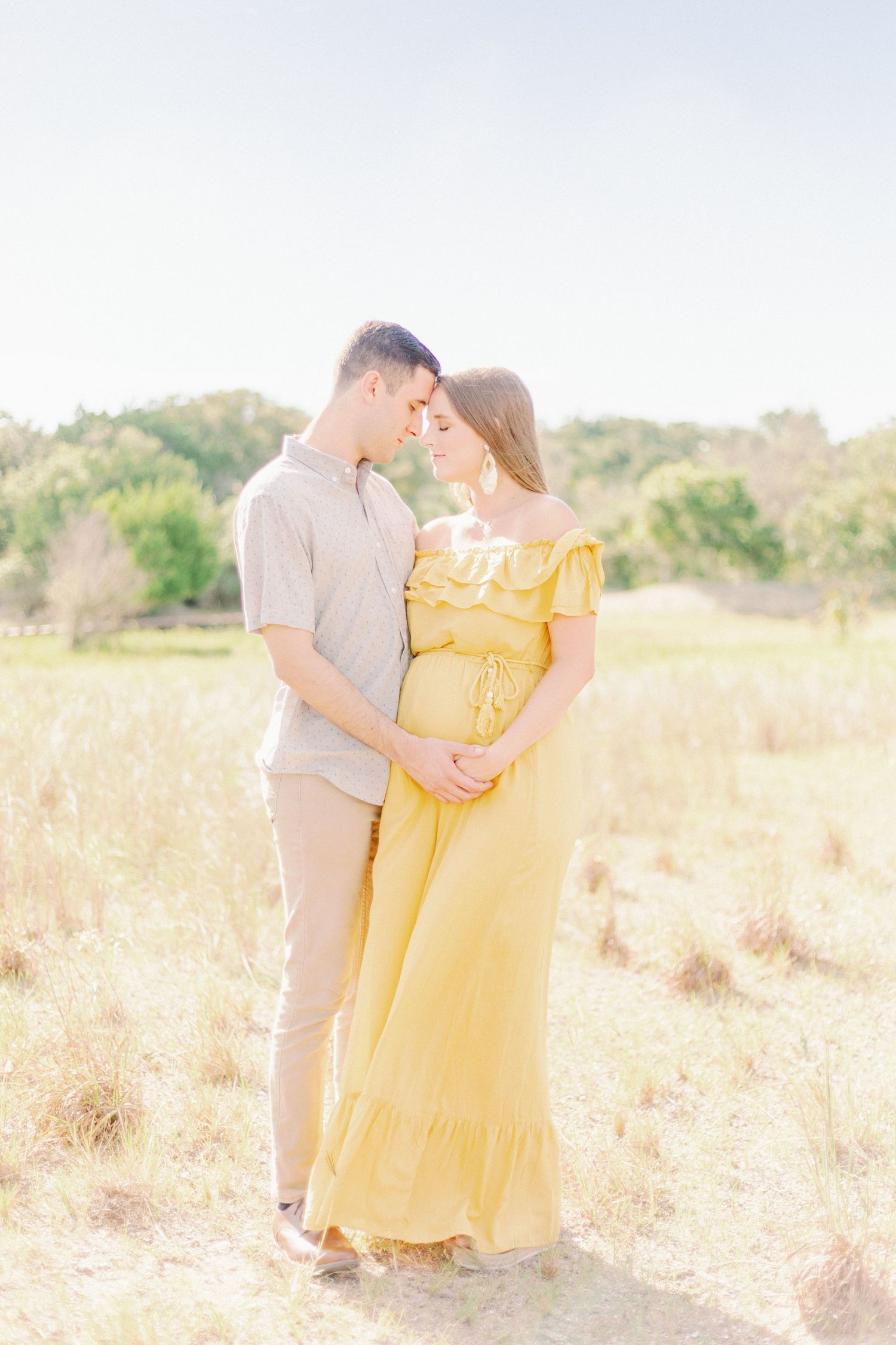 A couple embraces in a sunlit field with golden grass and soft natural lighting as they hold hands beneath the expecting mama's baby bump.