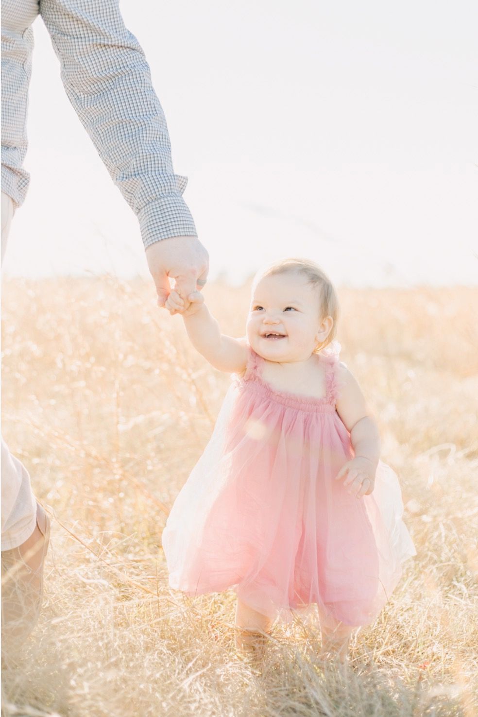 A small girl in a pink dress smiling while holding hands with someone in a Charleston, South Carolina field during golden hour.