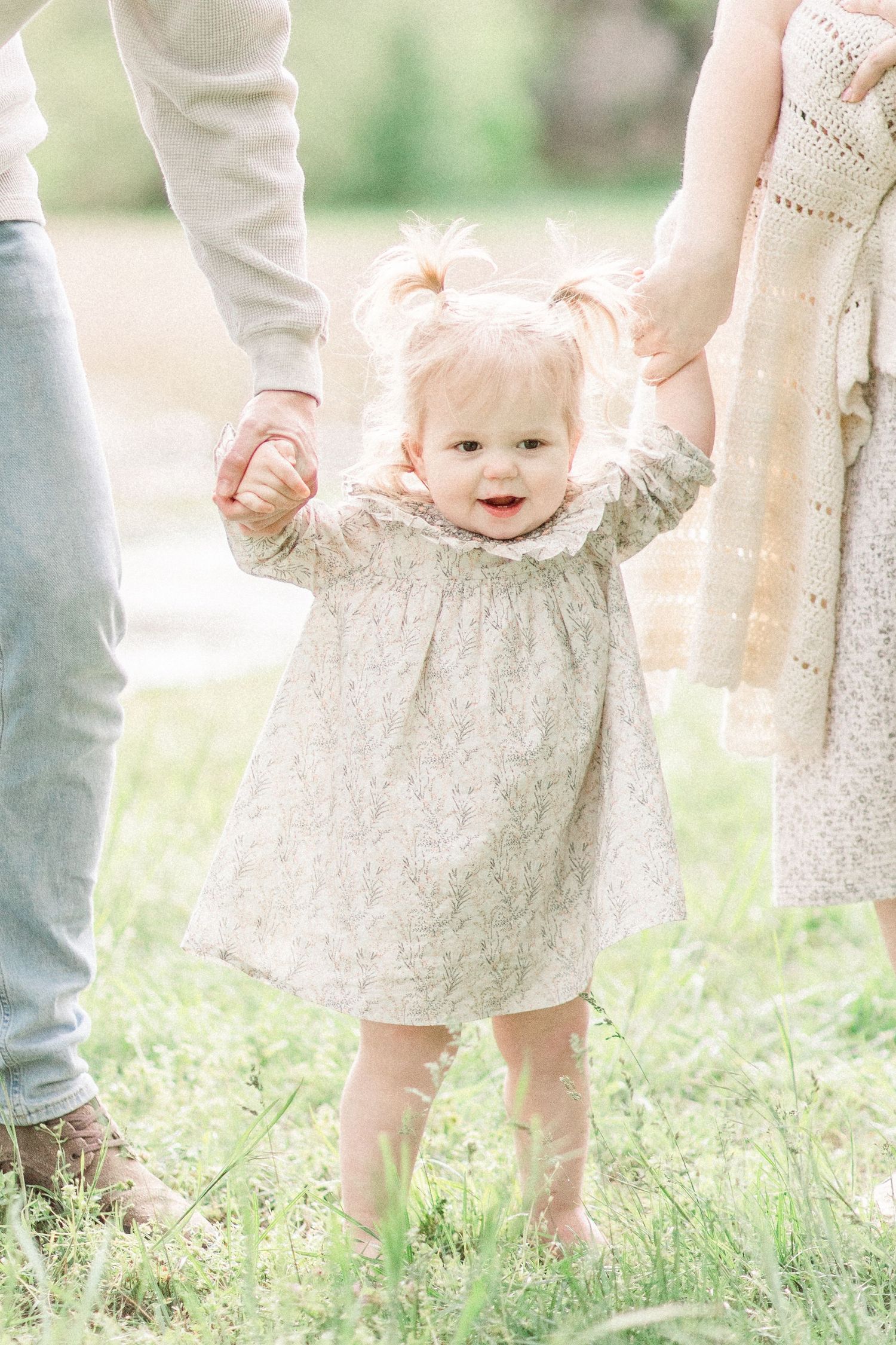 Small child in a lace dress holds hands with adults while standing in sunlit grass field in Springfield, Missouri.