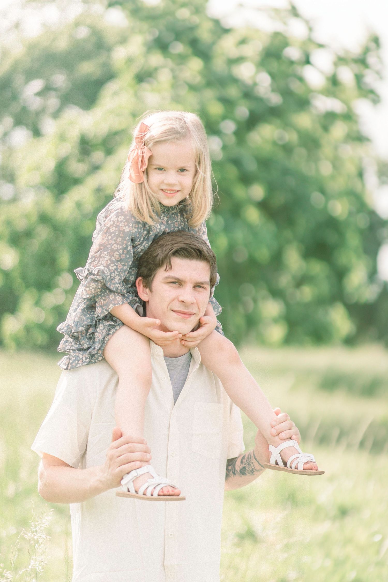 A Springfield, Missouri father gives a shoulder ride to a child in a floral dress on a sunny day in a grassy field.