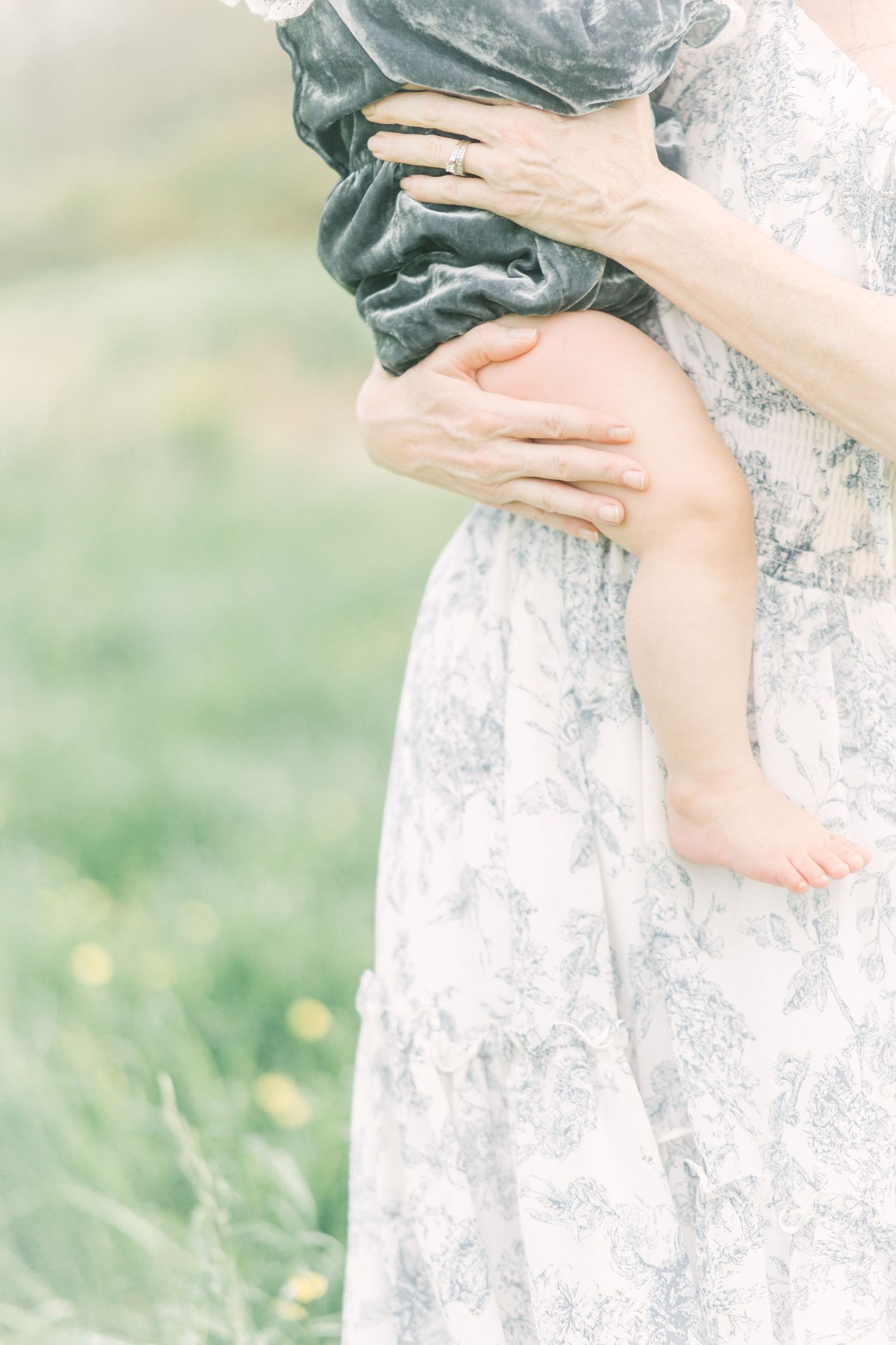 Gentle hands hold a baby's bare leg against a flowy white floral dress in a dreamy outdoor setting in Springfield, Missouri family session.
