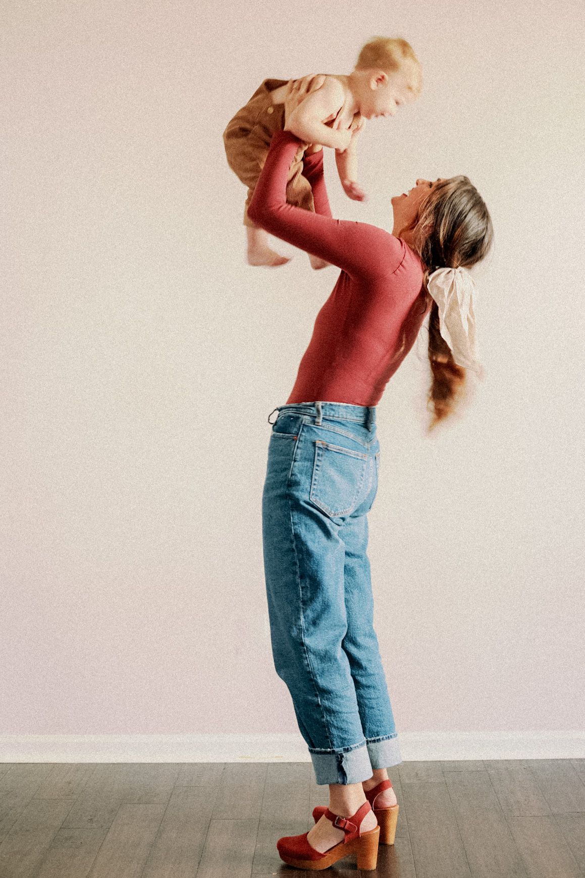 A mom in a red sweater and jeans lifts and plays with a small toddler in a beige outfit against a cream wall in Marshfield, MO.
