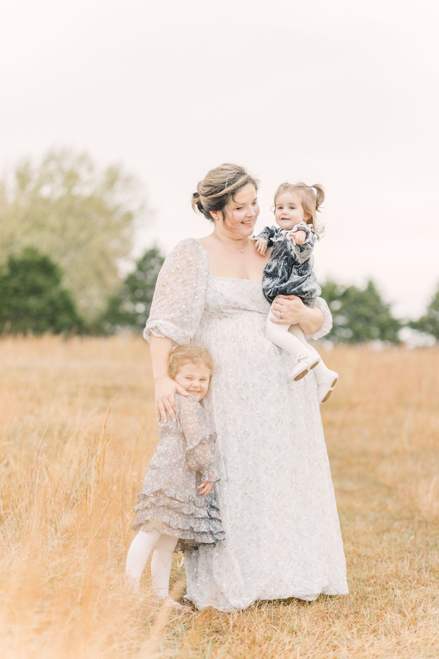 Pregnant woman in white lace gown holds toddler while young girl leans against her in golden field.
