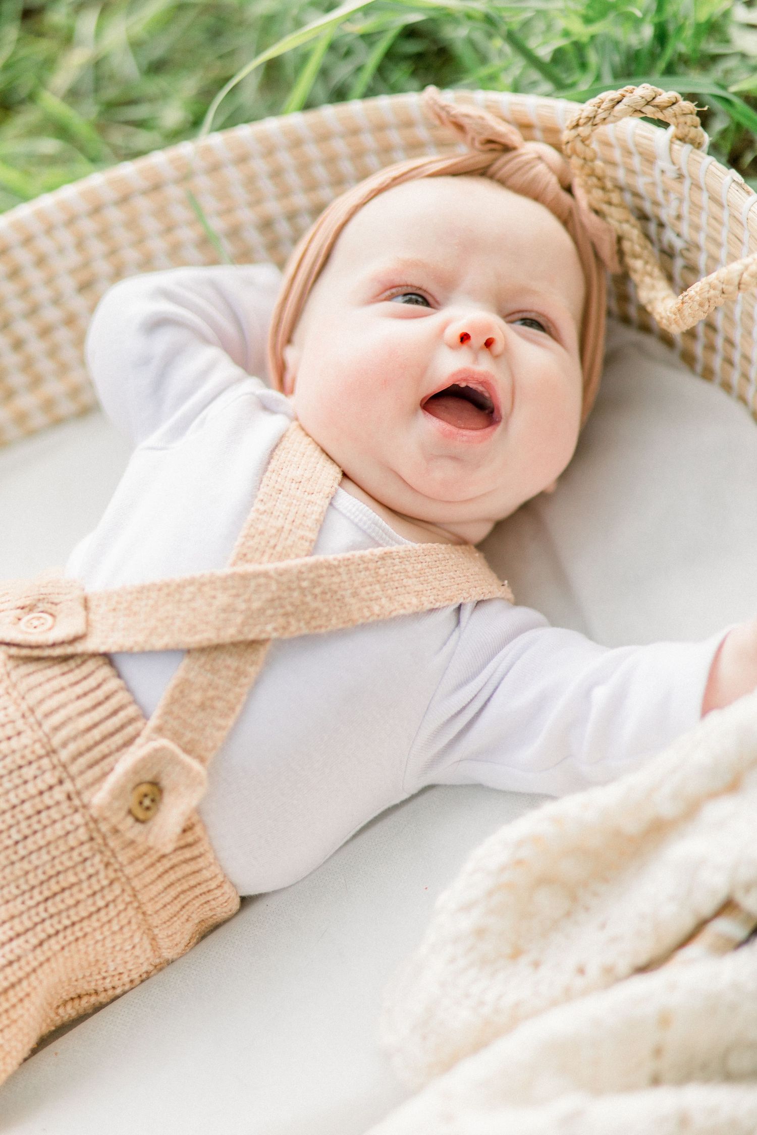 A baby wearing a knit beige overalls and white shirt lays in a Moses basket in Springfield, Missouri, smiling with joy.