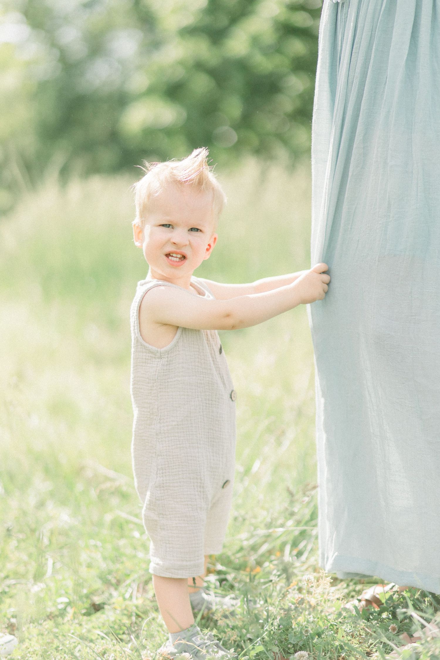 A toddler in a beige romper stands in a green meadow holding onto light blue fabric on a sunny day in Springfield, Missouri.