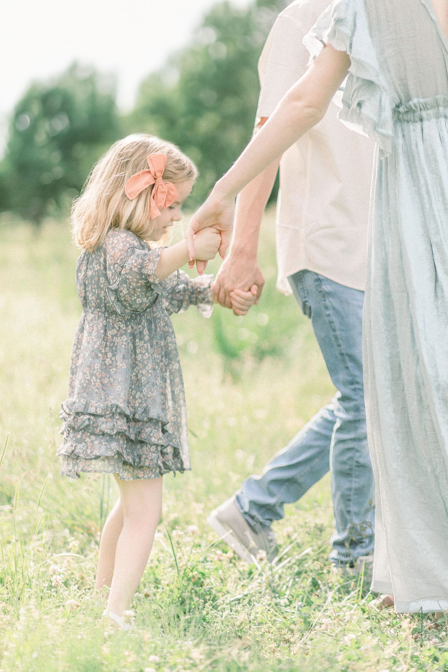 Young girl in a floral dress holds hands with her parents in a sunny Fellows Lake meadow with wildflowers in Springfield, Missouri.
