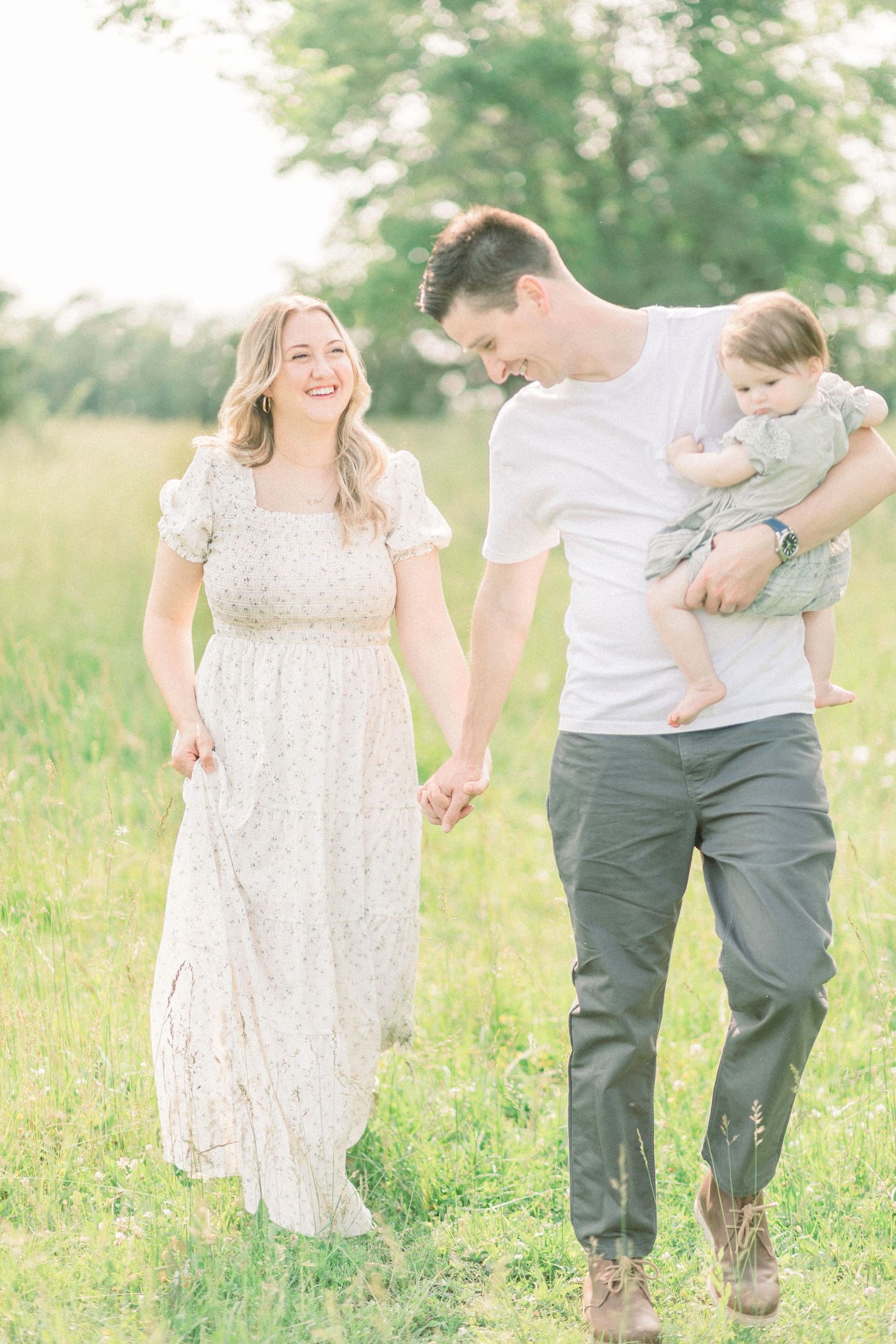 A happy young family walks through a sunlit green field, mother in floral dress, father holding baby.