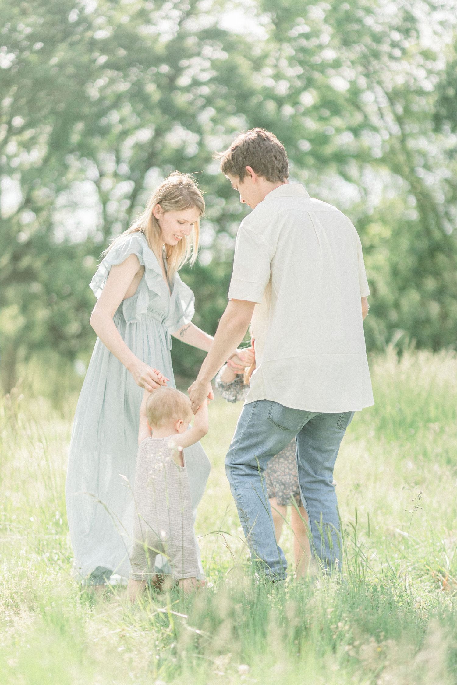 A family holds hands in a dreamy summer meadow with soft sunlight filtering through trees in the background in Springfield, Missouri.