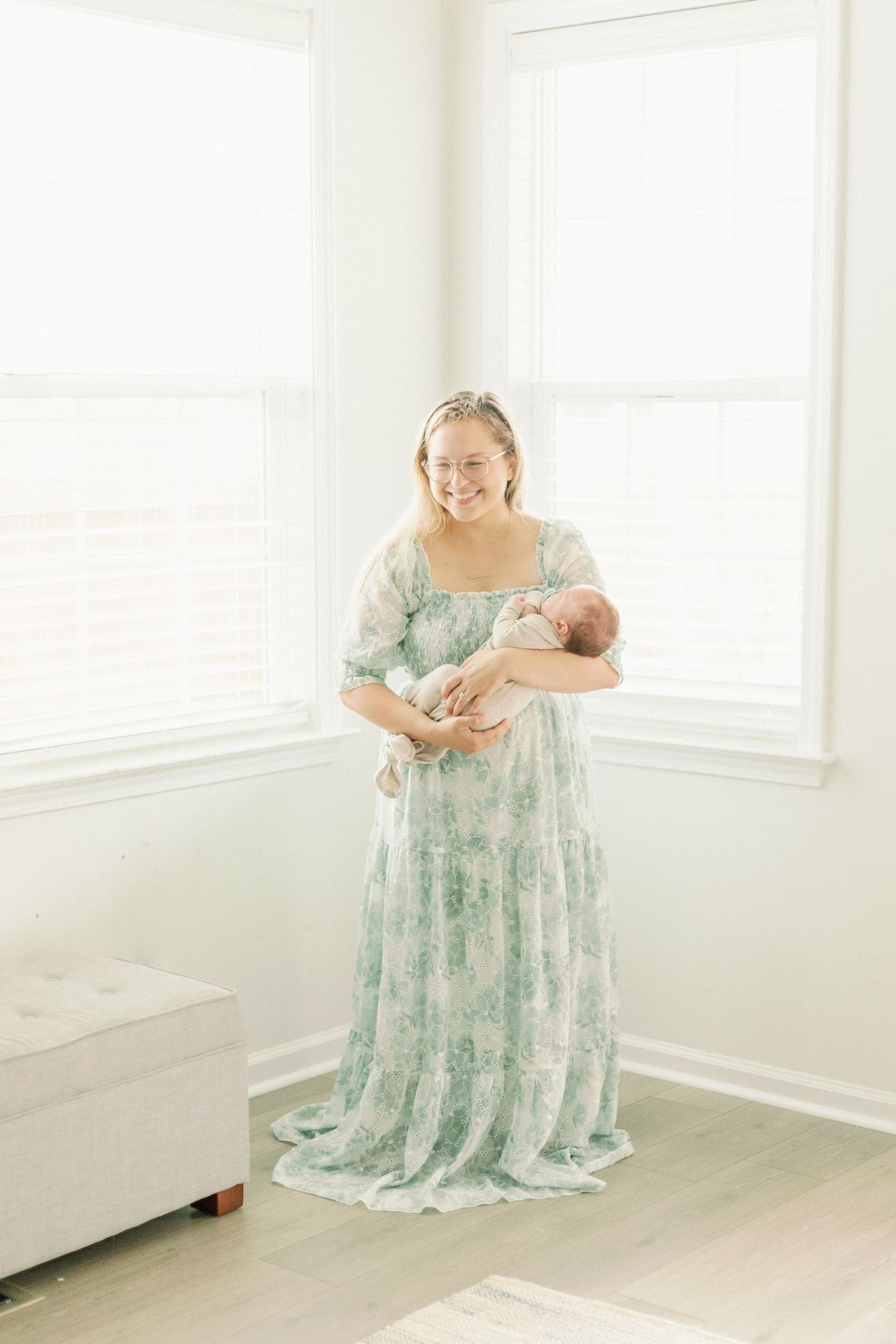Smiling mother in floral mint dress holds newborn baby in bright white room during lifestyle newborn photography session.