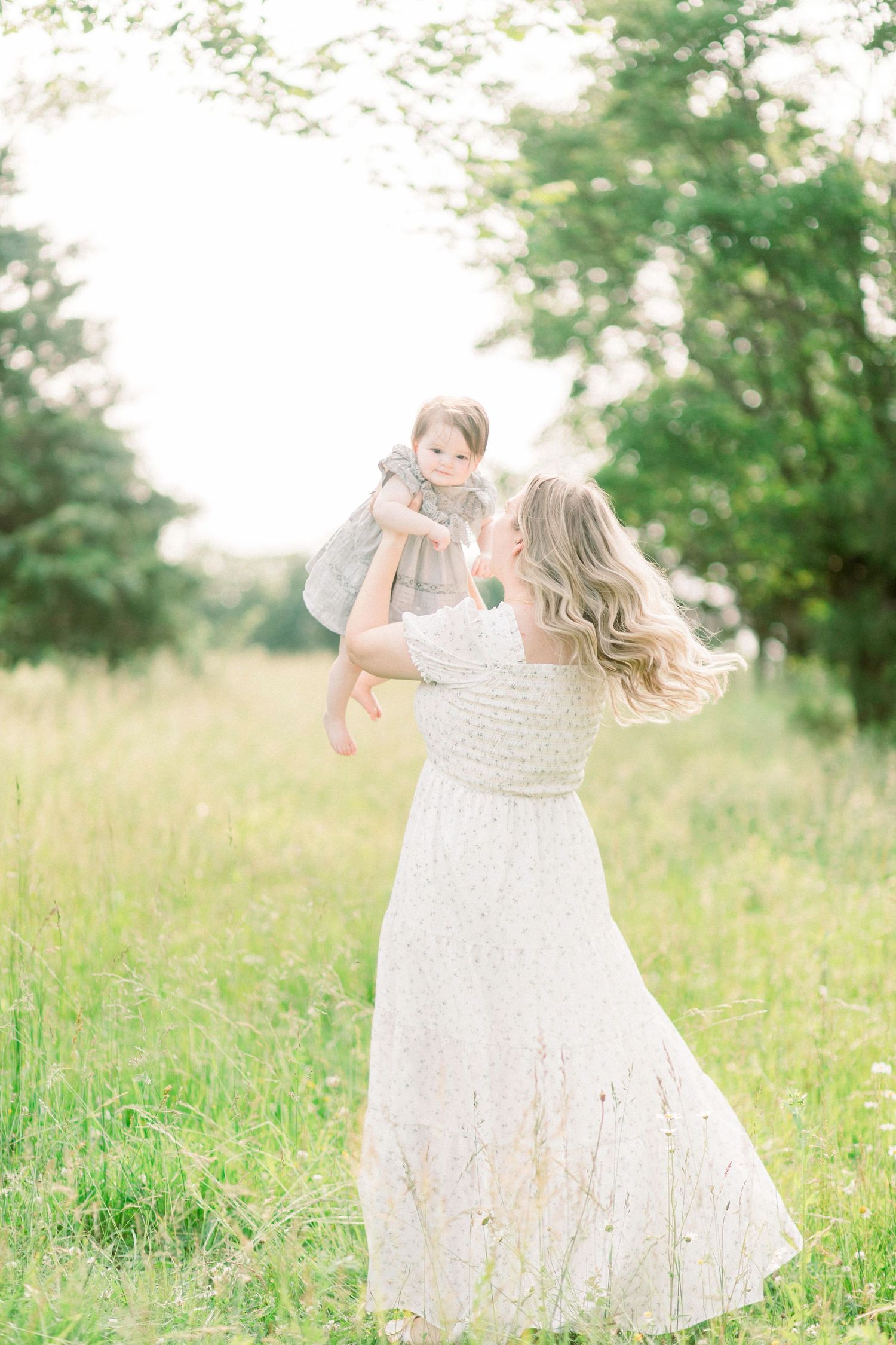 A woman in a flowing white lace dress lifts a small child in a sunny meadow with trees in the background in Springfield, Missouri.