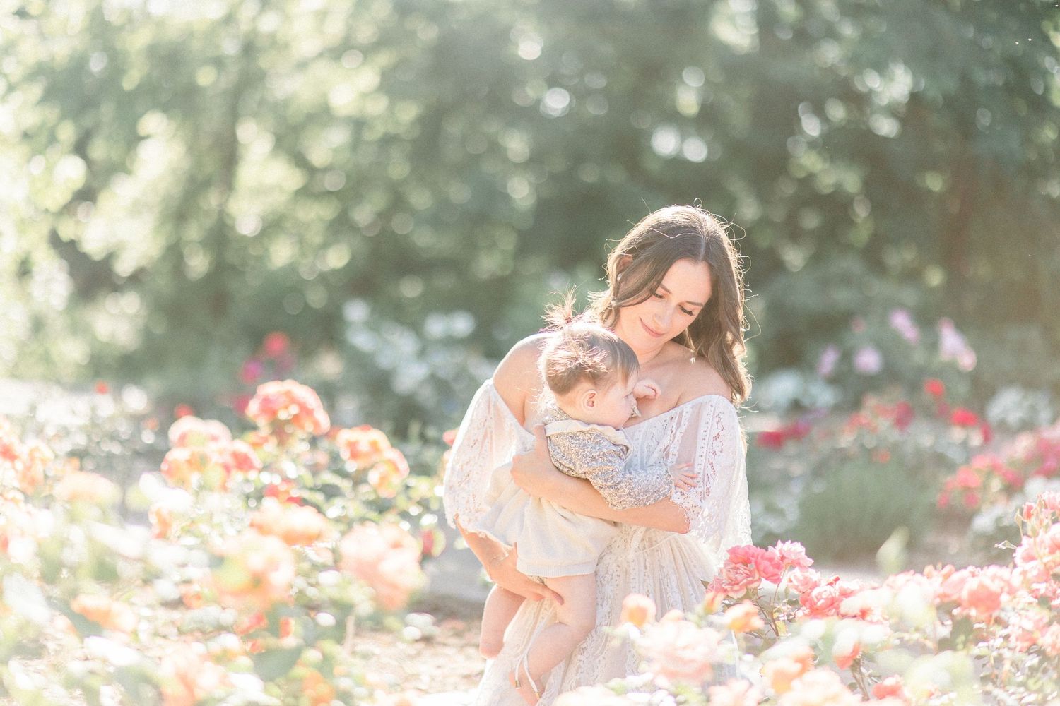 A mom in a lace dress holds her baby girl in a blooming rose garden on a sunny summer day in Nathaniel Greene Park in Springfield, MO.